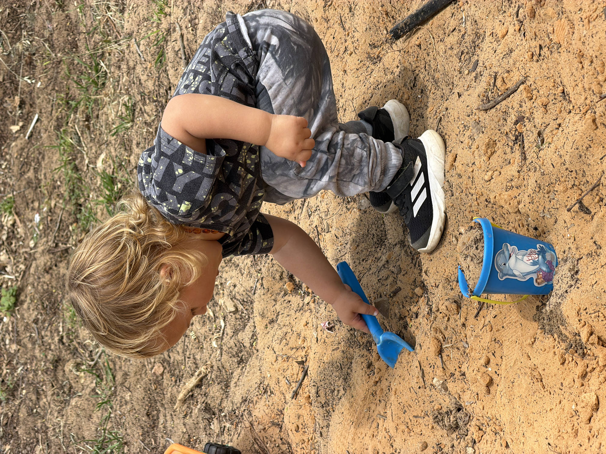 A young child with blonde hair, wearing a patterned gray t-shirt and gray patterned pants, is playing in a sand area. The child is bending over and holding a blue plastic toy shovel in their right hand, digging in the sand. A blue plastic bucket with a cartoon dolphin sticker is lying on its side in the sand to the right of the child. The child is wearing black and white Adidas sneakers. The surrounding environment appears to be an outdoor play area with a mixture of sand and soil. The location is Kiryat Bialik, Israel.