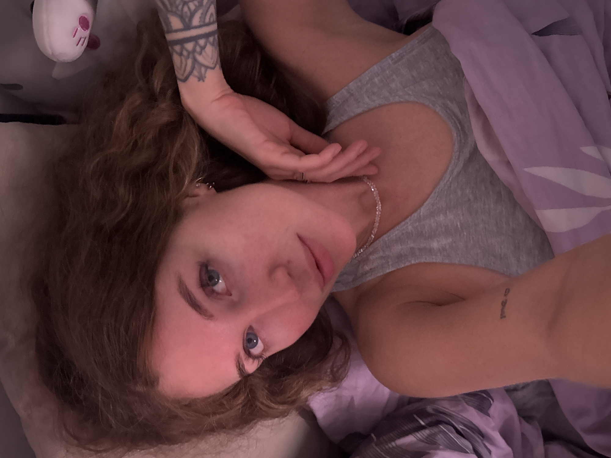 The image depicts an adult woman, appearing to be in the city of Chaikovsky, Russia, lying on her back in a bed and looking directly into the camera. Her reddish-brown, wavy hair is spread across a light-colored pillow. She has blue eyes, a fair complexion with some redness, and appears to be wearing minimal or no makeup. She is dressed in a grey tank top. Visible accessories include small hoop earrings on her left ear, a delicate beaded choker necklace, and a simple ring on her right ring finger. A tattoo featuring a geometric or mandala-like design is visible on her left forearm, and a small script tattoo is on her right wrist/forearm. Her right hand rests near her neck. She is partially covered by a purple duvet with a subtle white pattern. In the upper right background, a white plush toy with pink facial features, resembling a ghost, is visible. The scene is illuminated by dim, warm lighting, casting a soft, reddish hue.