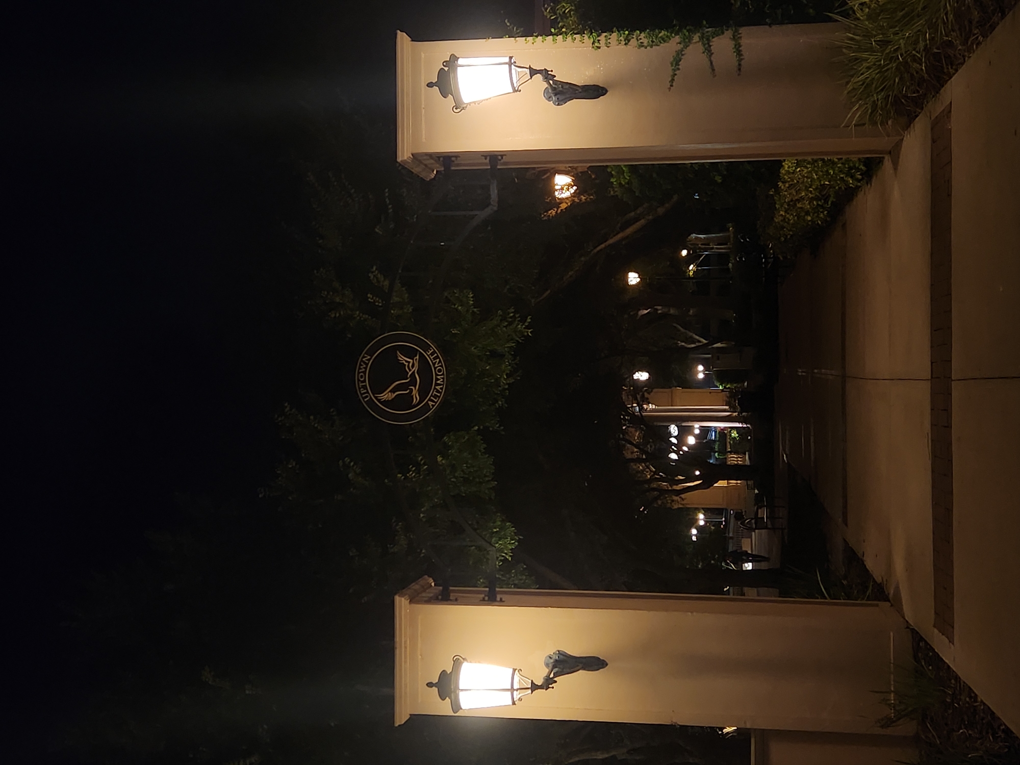 The image shows an entranceway at night, illuminated by ornate streetlights and wall-mounted lamps. An arched metal structure spans the pathway, bearing a circular emblem that reads "UPTOWN ALTAMONTE". Two large, cream-colored pillars flank the entrance, each adorned with a lantern. Beyond the entrance, a paved walkway stretches into the distance, lined with trees and bushes. Several other streetlights are visible further down the path, casting a warm glow on the scene. A person is faintly visible in the middle ground, walking away from the camera. The overall impression is one of a well-lit and inviting nighttime promenade.