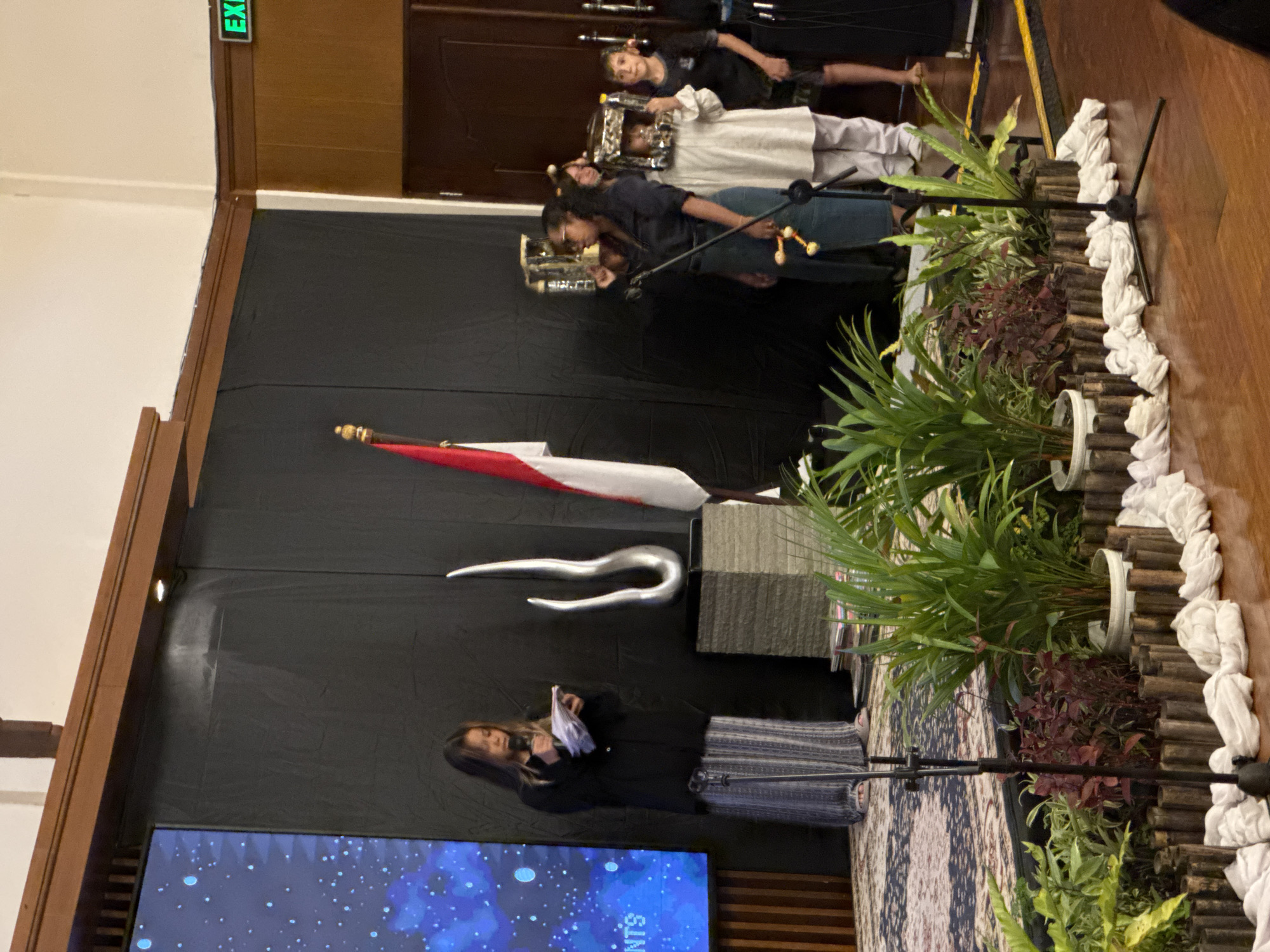 In an indoor venue, a woman stands at a podium, speaking into a microphone while holding papers, likely a script. Behind her, a tall, abstract silver sculpture sits on a stacked base. To her right, a flag with red and white horizontal stripes is visible. Several other individuals, including children wearing elaborate headpieces or costumes, are positioned further back. The background features a large black curtain and a screen displaying a blue, star-like pattern. The scene appears to be a formal event or presentation, possibly a cultural or educational gathering. An "EXIT" sign is visible in green lettering above a doorway on the right. The foreground is decorated with potted plants and a wooden border, with white fabric elements placed along the edge of the wooden floor. The overall setting suggests a public event space.