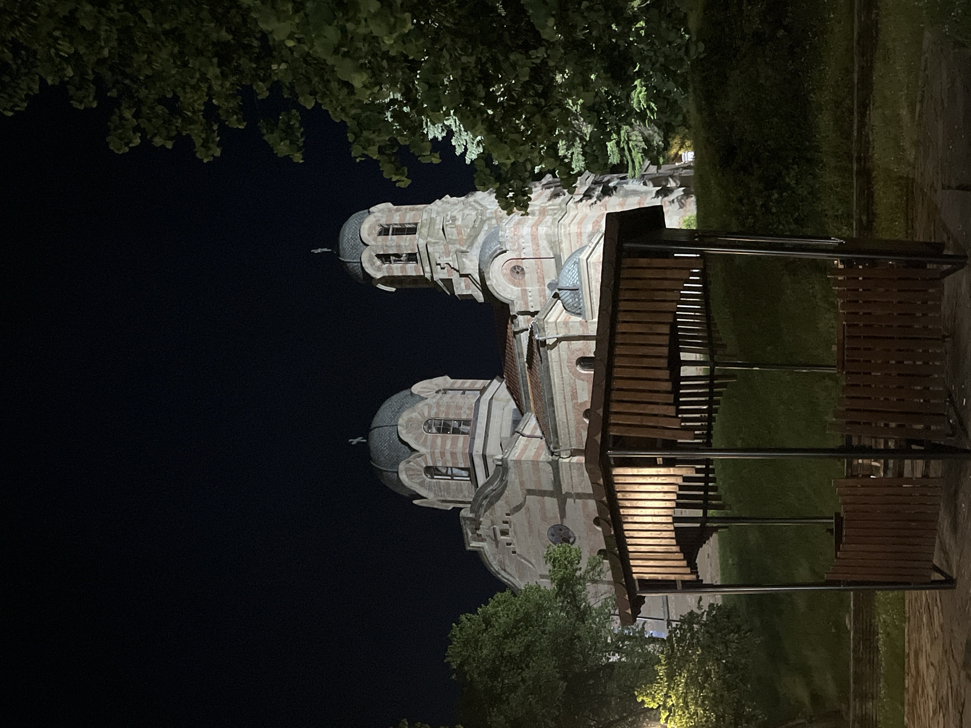 The image depicts the Church of Saints Peter and Paul in Veliki Preslav, Bulgaria, at night. The church, a prominent structure in the midground, features a distinctive striped facade composed of alternating bands of light-colored stone and reddish-brown brick. Two main domes are visible: a larger central dome to the left and a smaller dome to its right. A taller bell tower is situated on the right side of the church, also exhibiting the striped pattern and featuring arched openings. All visible domes and the bell tower are topped with Orthodox crosses.
In the foreground, a hexagonal wooden gazebo occupies the lower-middle portion of the frame. It has a dark, possibly metal, structural frame and walls constructed from vertical wooden slats. An internal light source illuminates the interior of the gazebo and the inner surfaces of its wooden slats, casting a warm glow. No individuals are present within or around the gazebo.
Trees with dark green foliage flank the church on both sides, with a dense cluster of trees and bushes partially obscuring the right side of the church and extending into the background. Another tree is visible on the far left. The ground immediately surrounding the gazebo appears to be paved with stones, transitioning to a grassy slope behind it. The sky above is dark, indicating nighttime, with no celestial bodies visible. Artificial lighting illuminates the church's facade and the gazebo, contrasting with the dark surrounding environment.