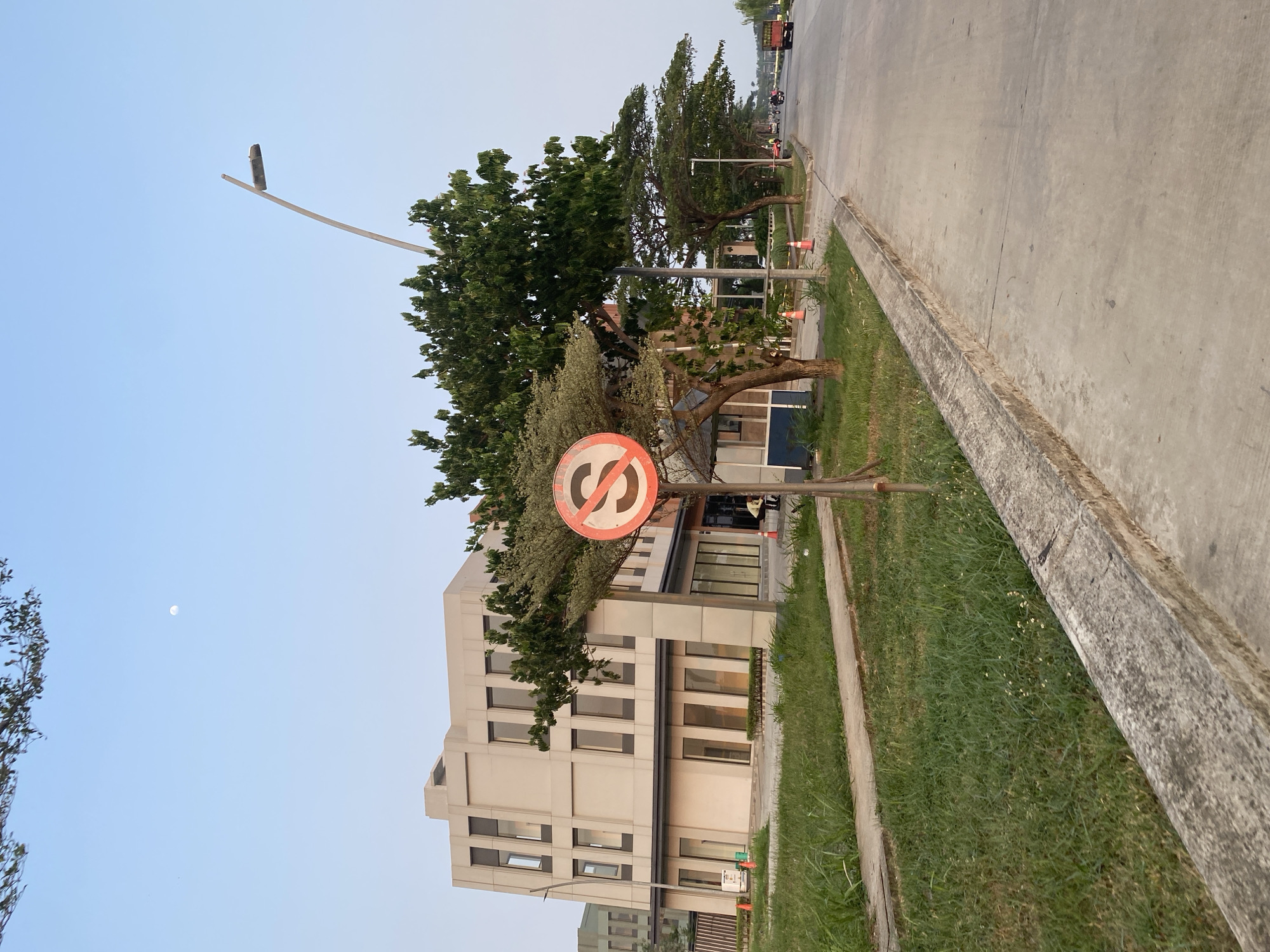 The image depicts a street scene with a modern building on the left and a road on the right, bordered by a grassy verge and a concrete curb. A circular traffic sign with a red border and a diagonal red line across a black "S" symbol is prominently displayed on a wooden post. Orange traffic cones are visible on the grass.
In the background, there are trees and a glimpse of other buildings. The sky is clear and pale blue, with a faint crescent moon visible. The lighting suggests it is either early morning or late afternoon. The overall scene appears to be in a developed urban or suburban area, with clean sidewalks and manicured grass. No specific activity or event is clearly depicted beyond the static elements of the environment. The image is oriented vertically. The location is East Jakarta, Indonesia.
