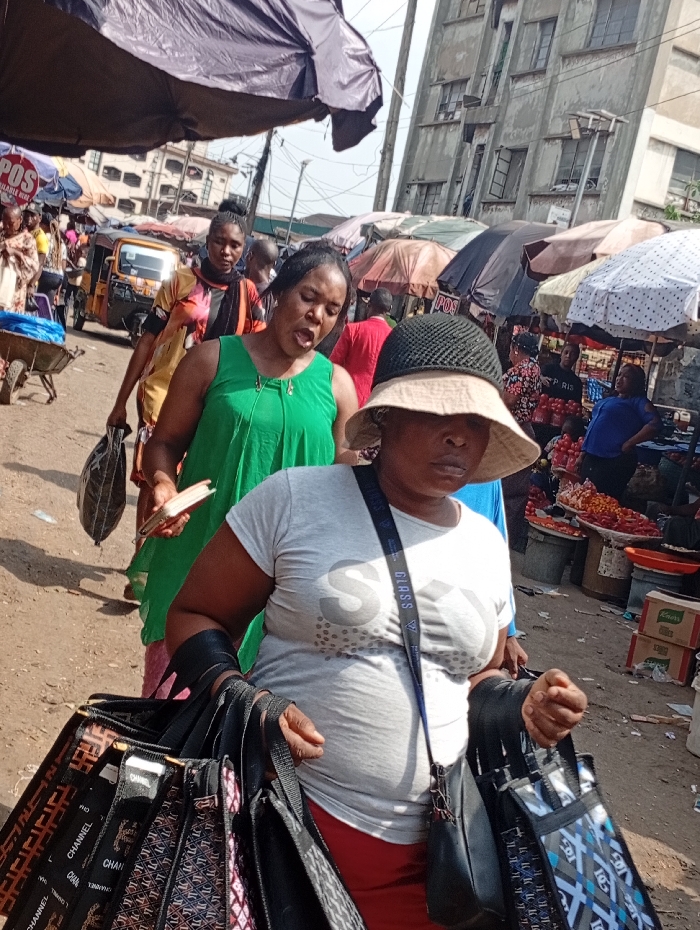 The image depicts a bustling street market in Onitsha, Nigeria, under daylight conditions. In the foreground, a woman in a light grey "GUESS" branded t-shirt, red lower garment, and a beige bucket hat with dark netting carries multiple black tote bags, some bearing "CHANNEL" branding. She also wears a black sling bag with a blue "GUESS" strap. Her right arm has a dark sleeve or brace. Behind her, another woman in a green sleeveless dress carries a light-colored object in her left hand and a dark bag in her right, with her mouth open. Further back, another woman in a patterned top walks along the unpaved, dusty ground.
The environment is characterized by numerous market stalls featuring various goods, including a display of red produce (likely tomatoes or peppers). Multiple market umbrellas and tarps provide shade. Two "POS" (Point of Sale) signs are visible on different stalls. In the midground, an orange auto-rickshaw (keke napep) and a wheelbarrow are present. The background features several multi-story, weathered concrete buildings with multiple windows, typical of urban structures in the region. Overhead, power lines are visible against a bright sky. The scene shows active pedestrian movement and commercial activity.