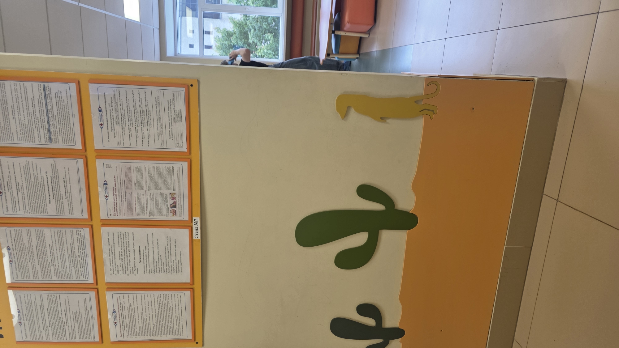 The image shows a wall with information posters and desert-themed decorations, likely in a public or institutional setting in Saint Petersburg, Russia.
On the left side, there's a wall-mounted board with multiple information posters in Russian, enclosed in yellow frames. The posters contain text, possibly related to rules, guidelines, or important notices. The text on one poster refers to "Прокуратури Красногвардейского района Санкт-Петербурга", suggesting the location within the Krasnogvardeysky district of Saint Petersburg.
To the right of the board is a section of the wall decorated with desert-themed elements, including a light-yellow sand-colored area and green cactus silhouettes. The background wall color is off-white.
In the background, there's a person lying down near a window, possibly using a phone. There's also a glimpse of furniture, including a chair.
The overall environment appears to be a hallway or waiting area, with a tiled floor visible.