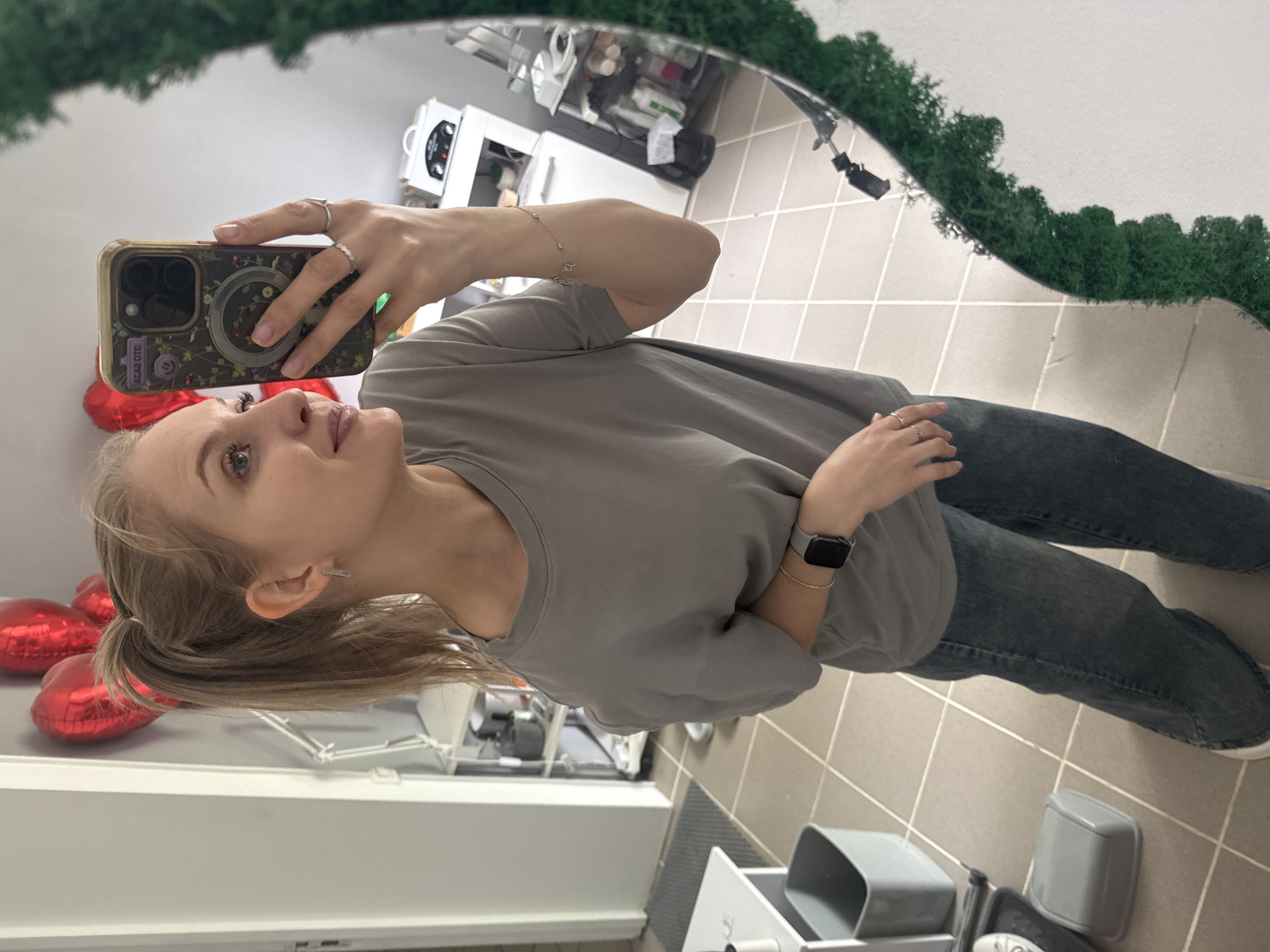 A woman with blonde hair pulled back in a ponytail is taking a selfie in a mirror. She is wearing a gray t-shirt, blue jeans, a watch on her left wrist, and rings on her fingers. The mirror has green moss trim. Behind her, there are red heart-shaped balloons, white cabinets, and gray tiles on the floor. The location appears to be a bathroom or dressing room. It is not possible to determine the exact location within Izhevsk, Russia from the image.