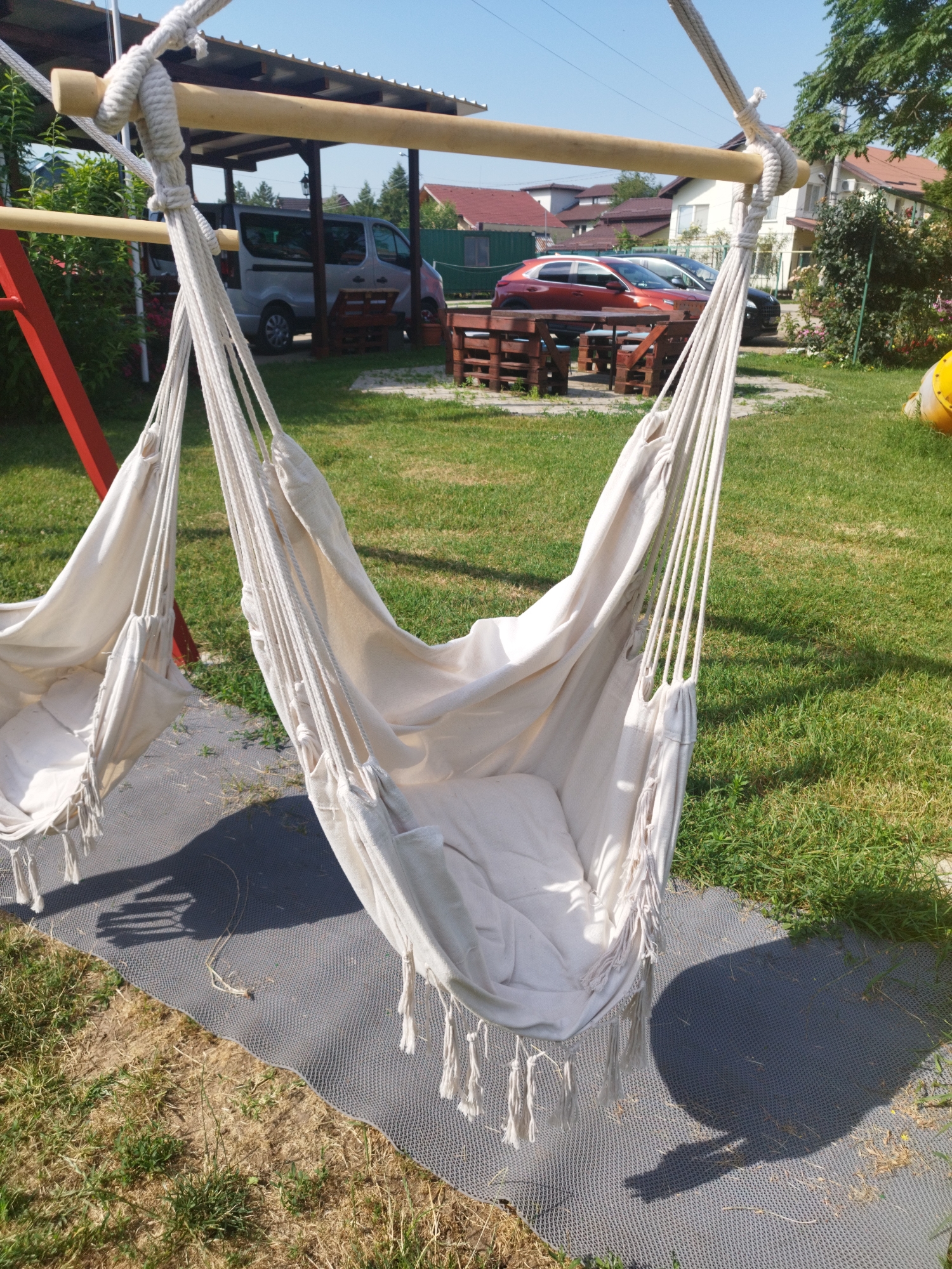 The image depicts a sunny day in a residential area of Bucharest, Romania. In the foreground, two cream-colored fabric hammocks hang from a wooden structure supported by red metal legs. The hammocks are made of woven material with decorative tassels at the bottom. One hammock is closer to the camera and appears to be empty, while the other is partially visible to the left.
The ground beneath the hammocks is covered by a dark grey textured mat, with dry grass and patches of green grass visible around it. In the background, a covered parking area with a corrugated metal roof is visible, sheltering a silver van. Beyond the parking area, several houses with red-tiled roofs and trees are seen under a clear blue sky. A red car is parked on the street in front of one of the houses, and further down the street, another dark-colored car is visible. The overall scene suggests a residential backyard or a communal outdoor space.