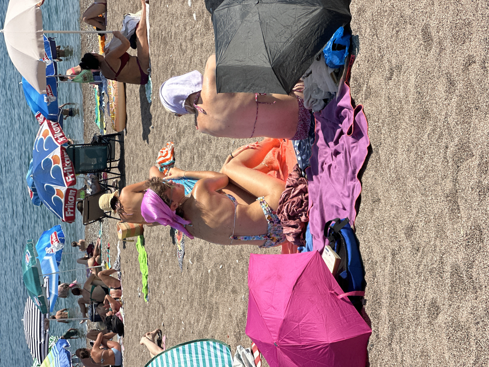 In Boreti, Montenegro, on a crowded beach, a group of people relax under colorful umbrellas. In the foreground, a bright pink umbrella is open, casting a shadow on a blue backpack and a book. To the right, a woman in a purple bikini sits under a black umbrella. Behind her, another woman in a floral bikini and a pink headscarf is partially visible. Several other individuals are scattered across the pebbled beach, some sunbathing on towels and others wading in the calm, blue water. Various beach umbrellas, including blue ones with "Frikotherm" branding, dot the scene, providing shade from the bright sun. The overall atmosphere is one of leisure and summer vacation.