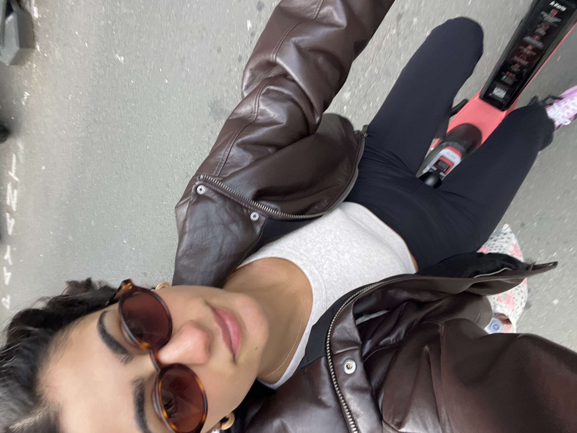 A woman wearing a brown leather jacket, white tank top, and black leggings is riding a pink electric scooter on a city street. She is wearing sunglasses. Her hair is dark. The scooter has controls and labels in English. The location is likely Paris, France, based on the scooter branding.