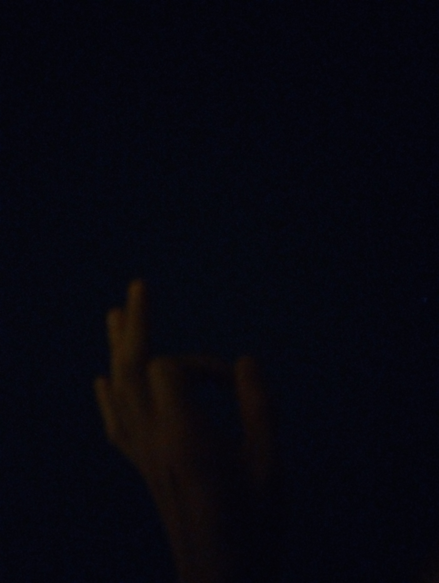 A close-up, dimly lit photograph shows a portion of a human hand with fingers extended. The background is entirely dark blue, suggesting a low-light environment. The hand appears to be positioned in the lower center of the frame, with the fingers pointing upwards. The lighting is such that only the outlines and some texture of the hand are discernible, giving it a somewhat ghostly appearance. No other objects, people, or environmental details are visible. The location is unknown.