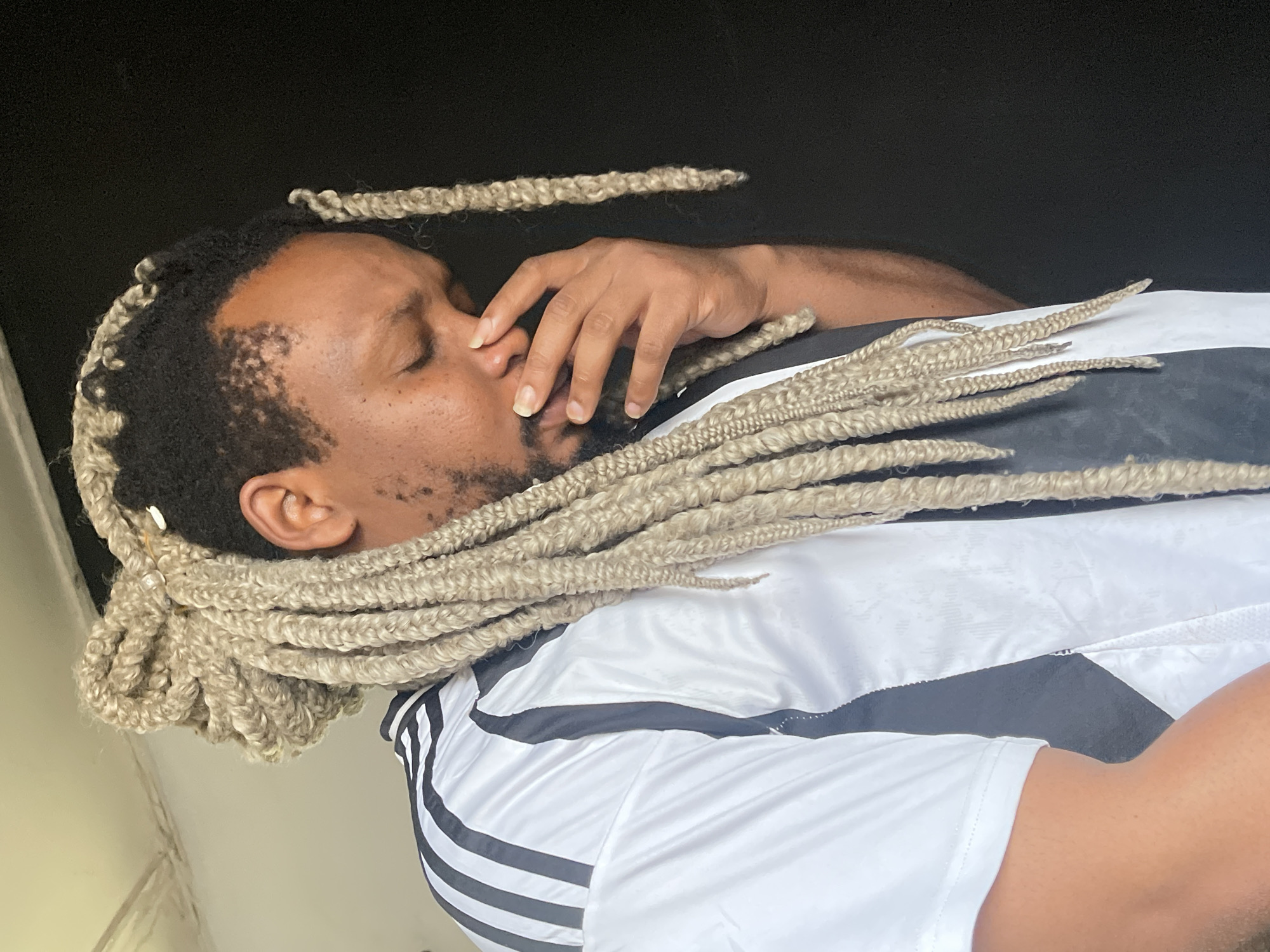 A man with blonde braids covering his hair is shown in this close-up portrait. He is wearing a white shirt with black stripes on the shoulders and down the sides. The man has his eyes closed and his hand is covering his nose and mouth, suggesting he is experiencing some discomfort or emotion. He is standing in front of a black background. The overall lighting suggests this is an indoor shot, possibly taken in a residential setting in Jos, Nigeria.