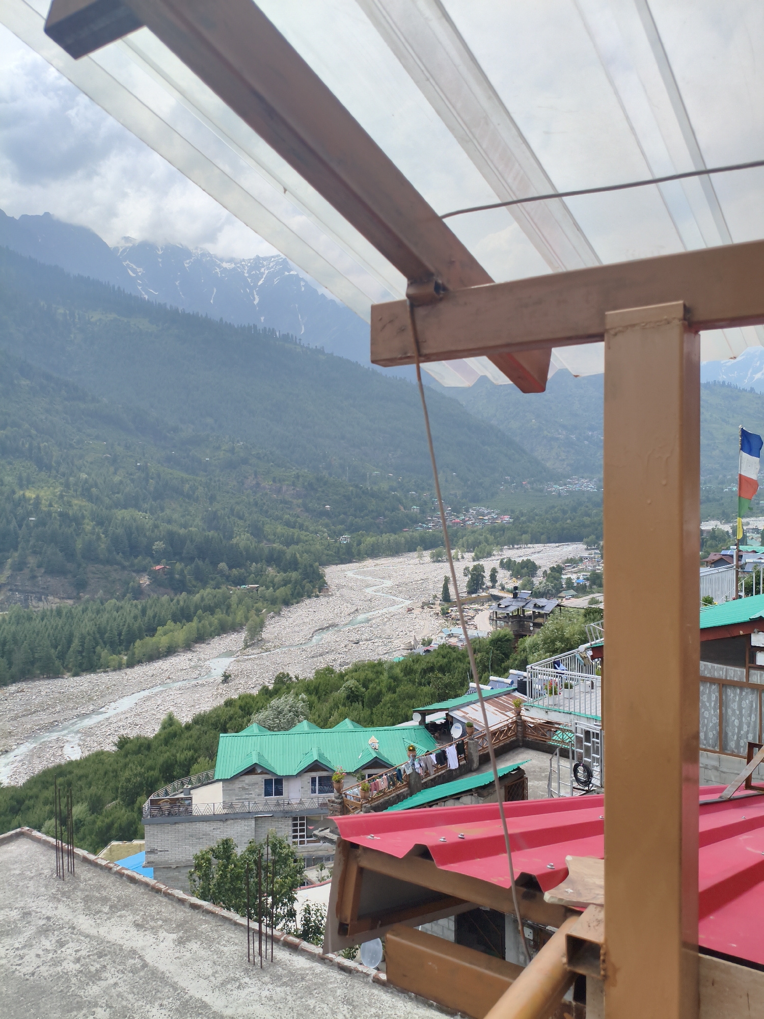 The image captures a scenic view from a vantage point overlooking the town of Vashisht, India, with a river valley and snow-capped mountains in the background. In the foreground, a rooftop area is visible, featuring a concrete surface with rebar protruding from it, suggesting ongoing construction or a weathered structure. A red metal roof is also prominent, with visible screws holding it in place.
The midground reveals a tiered settlement of buildings, many adorned with vibrant green metal roofs, characteristic of the region's architecture. Laundry hangs on clotheslines outside some of the structures, indicating daily life. A French flag is visible on a flagpole, adding a touch of international presence to the scene. A wider view shows a rocky riverbed, with a clear stream flowing through it, bordered by lush green trees and dense foliage that blankets the slopes of the surrounding mountains. The majestic Himalayas, with snow dusting their peaks, form a dramatic backdrop, partially obscured by clouds. The overall impression is one of a peaceful, mountainous settlement nestled within a natural landscape.