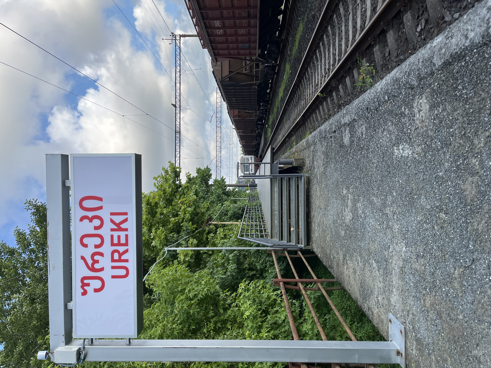 The image shows a train station platform in Ureki, Georgia. A sign with the name "UREKI" written in Georgian script and Latin letters is prominently displayed. The platform is made of concrete and gravel, with a metal staircase leading up to a walkway. To the right of the platform, there are train tracks with several freight cars. Lush green trees line the left side of the platform, and a cloudy sky is visible in the background. The scene appears to be a typical day at a provincial train station.