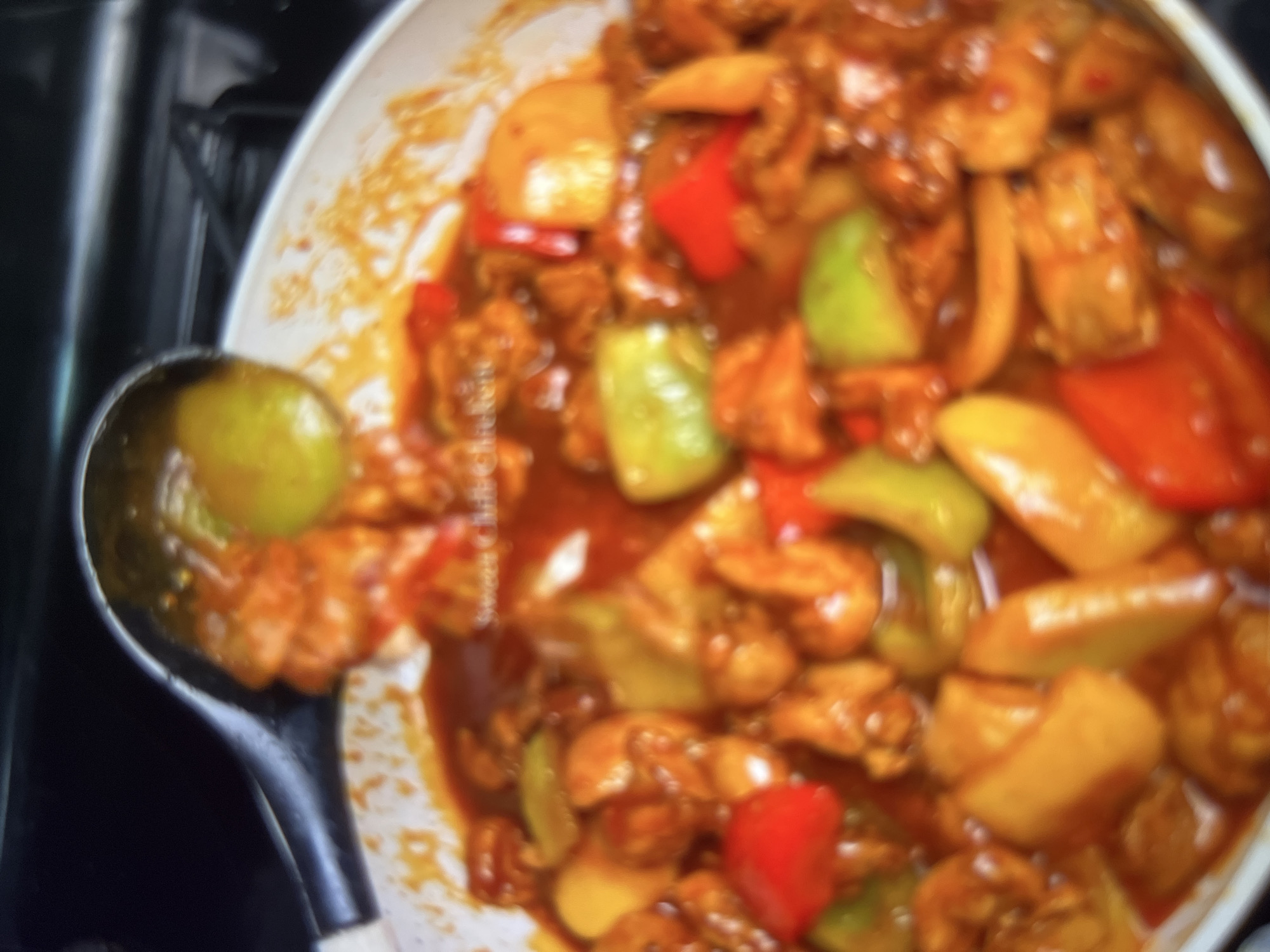 The image shows a close-up, blurry view of a dish of sweet chili chicken with peppers and onions in a white pan. A serving spoon is visible on the left side of the image, scooping up some of the food. The dish is a vibrant orange-red color from the sauce. There are no discernible landmarks or environmental details that can be used to determine a specific location in Abuja, Nigeria.