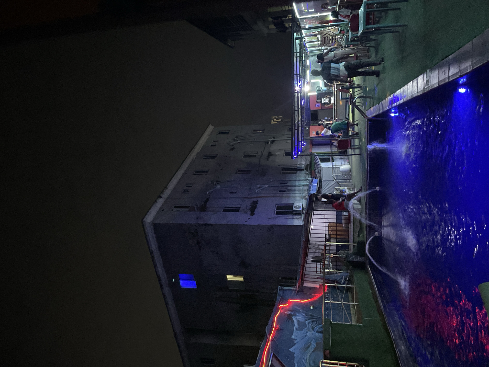 The image depicts an outdoor, elevated social area at night, likely a rooftop lounge or restaurant, in Ado, Nigeria. The scene is dominated by a rectangular swimming pool illuminated with vibrant blue lights, from which multiple jets of water arc into the air, creating a dynamic visual effect. The water also reflects a reddish glow from nearby string lights.
Adjacent to the pool, there is a seating area covered with green artificial turf, furnished with tables and chairs where several people are gathered. Some individuals are standing near the pool's edge, while others are seated and socializing. The atmosphere appears lively, highlighted by the various light sources.
In the background stands a tall, multi-story building, appearing somewhat weathered with a dark grey facade, and several windows. Most windows are dark, but a few emit a soft white or yellow light, and one prominent window on a lower floor glows distinctly blue. An air conditioning unit is visible on the building's exterior.
Further elements include metal railings, general overhead lighting illuminating the seating areas, and red string lights adorning a lower wall or railing, casting reflections into the pool. The sky is completely dark, indicating a late evening or night setting. No visible text is clearly legible within the image.