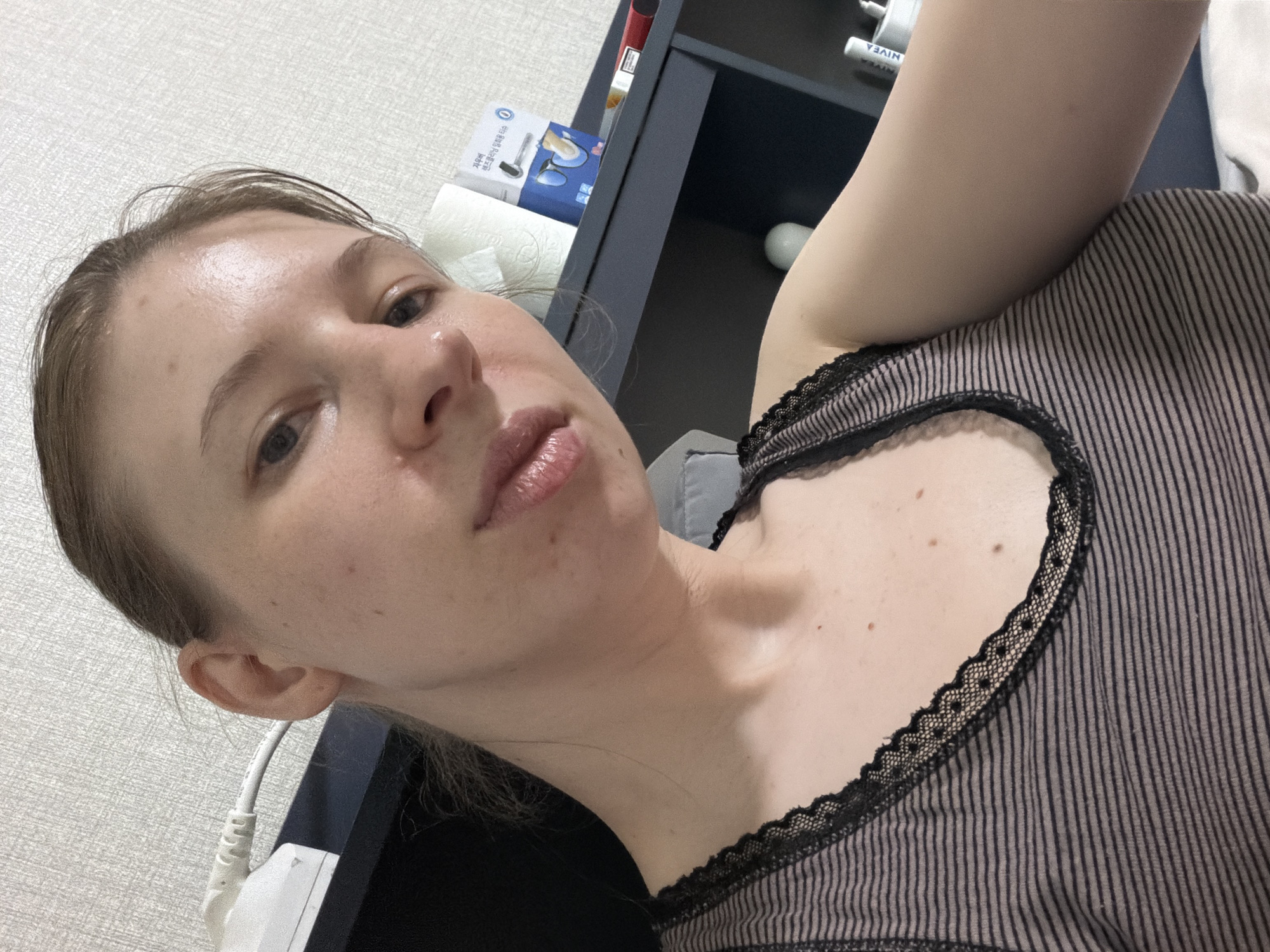 A young Caucasian woman with light skin and light brown hair pulled back is taking a selfie in a dimly lit room. She is wearing a striped tank top with black lace trim. Her face is turned towards the camera, with her eyes open and looking slightly to the left. Her lips are slightly parted, revealing a hint of teeth. The background consists of a white textured wall, a dark shelf with various items including a roll of toilet paper, a blue box with Korean writing and an image of contact lens cleaning supplies, a cigarette pack, and a white bottle. To the left of the woman, a white electrical outlet with a cord is visible. The image was taken in Dongducheon-si, South Korea.