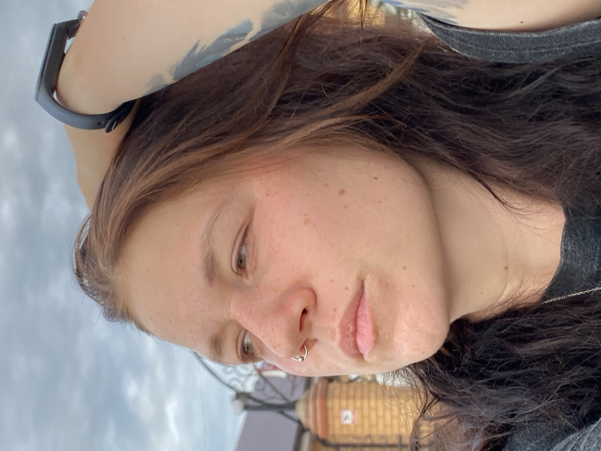 The image is a close-up, outdoor, waist-up shot of a young Caucasian woman with brown hair and freckles. She is looking to her left with her right arm raised, hand in her hair, and wearing a dark gray t-shirt and a black fitness tracker on her wrist. A septum piercing is visible in her nose. In the background, there is a cloudy sky and the side of a brick building with a black metal fence. The location is identified as Sergeevka, Russia.