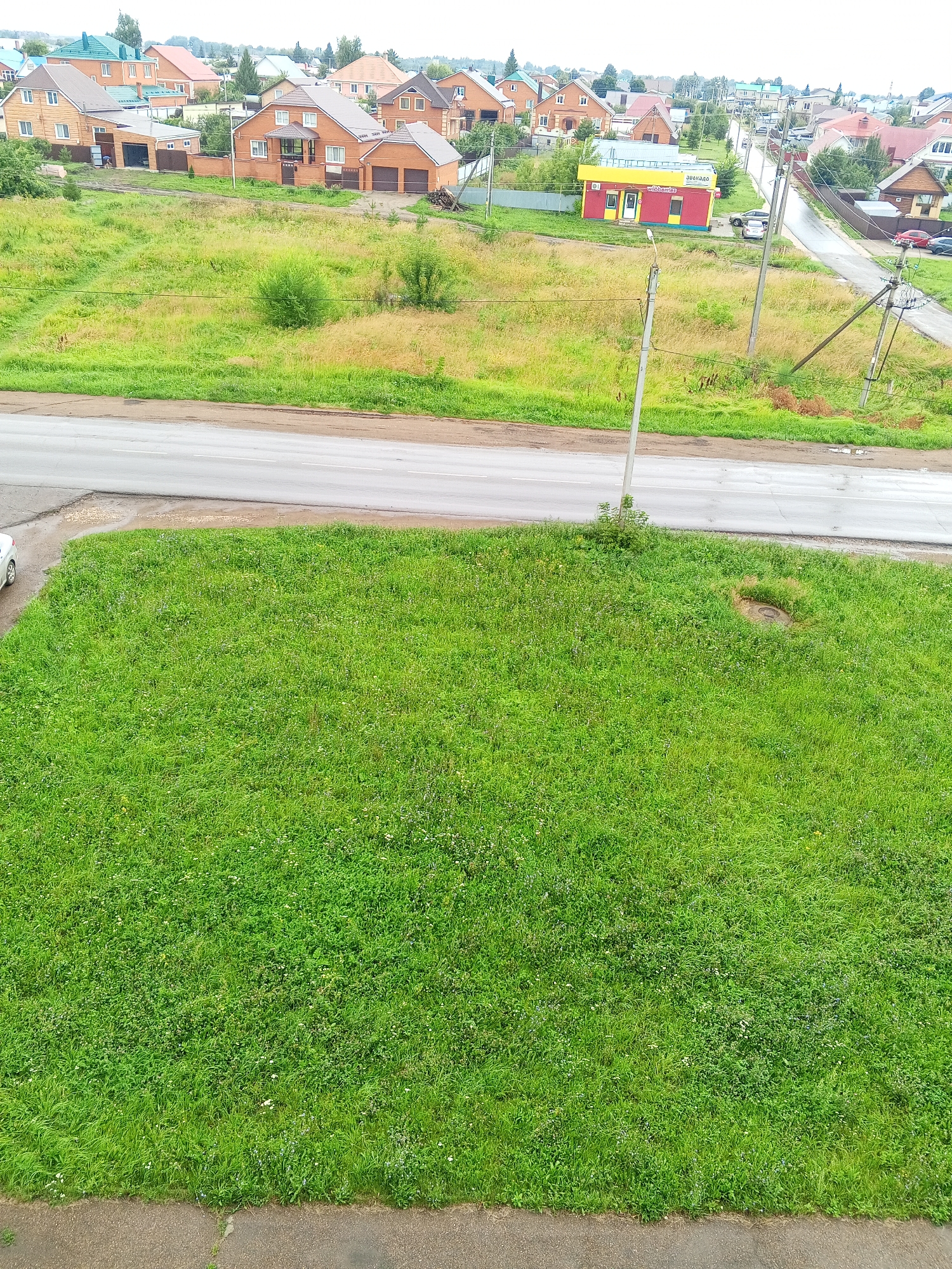 The image displays a suburban area in Chistopol, Russia, characterized by a mix of residential houses and open green spaces. In the foreground, a large expanse of well-maintained green grass dominates the frame, with a paved path or sidewalk along the bottom edge. A gray asphalt road runs horizontally across the middle ground, separating the foreground lawn from a field of taller, mixed-color grasses and brush. Beyond this field, a cluster of brick houses with red and orange roofs are visible, some featuring garages and fences. Further in the background, a two-story building with a bright yellow and red facade, appearing to be a small shop or kiosk, stands next to the road. Power lines and poles are scattered throughout the scene, indicating infrastructure within the area. The sky is overcast, suggesting a cloudy day. No people are clearly visible in the image, but a white car is partially seen on the left edge of the road. A circular manhole cover is embedded in the grassy area in the middle right of the frame.