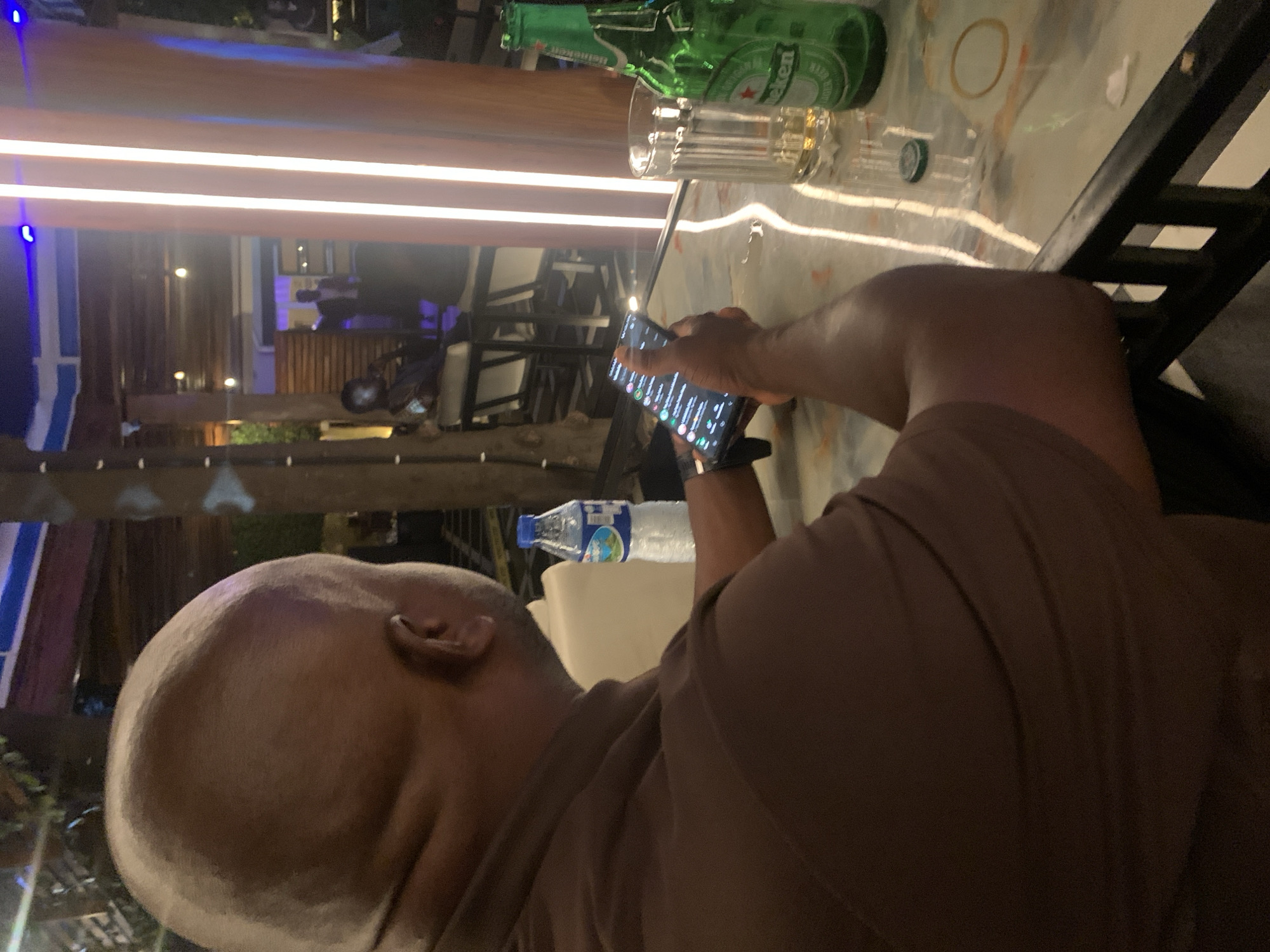 The image depicts a man, viewed from behind and to the side, sitting at a table in what appears to be an outdoor dining or lounge area. He is wearing a brown t-shirt and is holding a smartphone in his right hand, looking at its screen. On the table in front of him are a bottle of water, a glass, and a green bottle of Heineken beer. The background suggests an evening setting with ambient lighting, including vertical illuminated pillars, and glimpses of other patrons and structures in the distance. The style of the decor and lighting is consistent with modern establishments found in cities like Abuja, Nigeria, which often feature open-air seating and contemporary design elements.