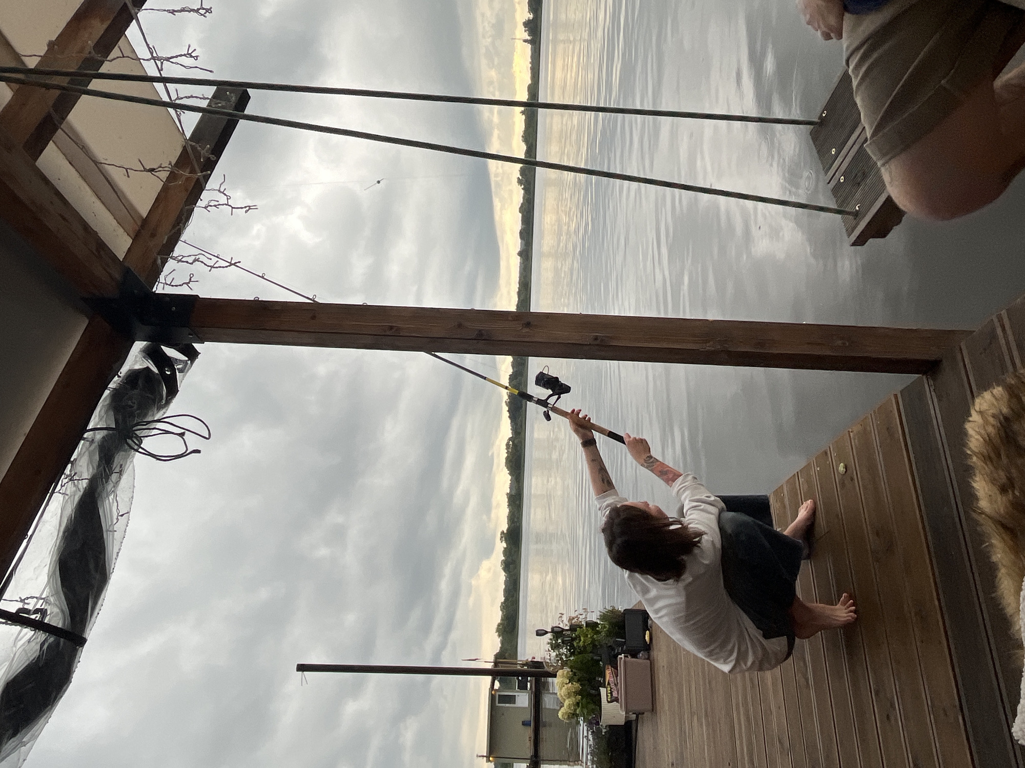 In Radchenko, Russia, a person is seen fishing from a wooden dock extending over a body of water under a cloudy sky. The individual, positioned in a squatting stance with their back to the camera, is holding a fishing rod cast out over the water. They are wearing a light-colored long-sleeved shirt and dark pants. To the right of the frame, a wooden swing hangs over the water. In the background, a small houseboat is visible on the water, and the distant shore is lined with trees. The dock's surface is made of wooden planks. The overall atmosphere suggests an overcast, tranquil day.