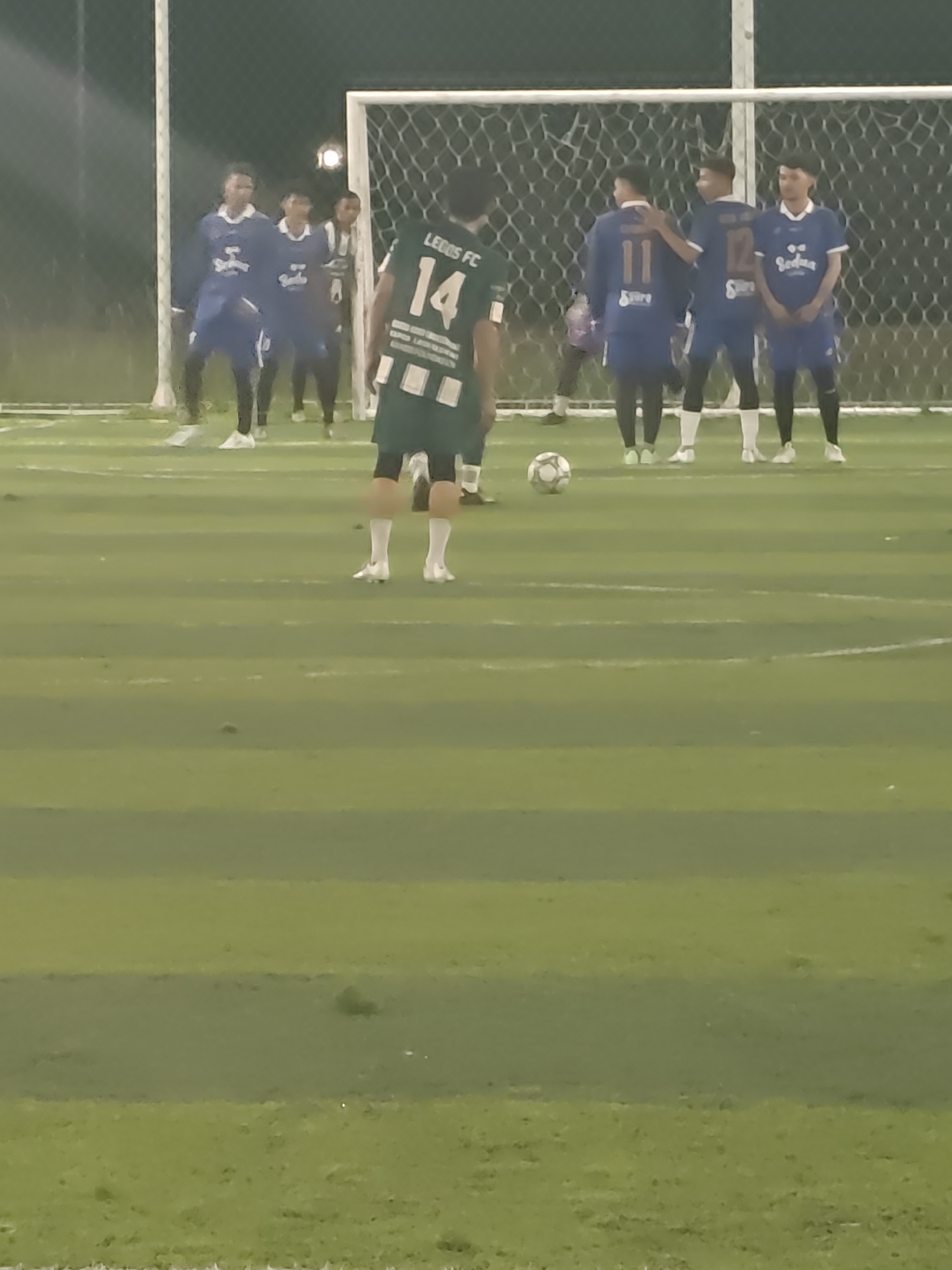 The scene depicts a soccer game on an artificial turf field, likely in Lhokseumawe, Indonesia, given the attire of the players, although this cannot be definitively stated. A soccer ball is present on the field. One player, wearing a green jersey with the number 14 and "Leeds FC" on the back, stands near the ball, apparently preparing to kick it. Across from him stand four players in blue jerseys. A soccer goal is behind the players. Additional players are in the background. The lighting suggests the game is taking place in the evening.