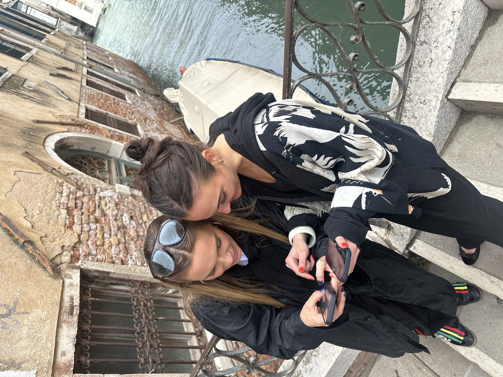 Two women are standing on a bridge in Venice, Italy, looking at a smartphone. The woman on the left is wearing sunglasses, a black coat, and black shoes with colorful stripes. The woman on the right is wearing a black and white jacket, black pants, and black shoes. They are standing on a stone bridge with a metal railing overlooking a canal. A boat is docked on the canal. Behind the women is a weathered building with brick and stone details. The building has several windows with iron bars. The woman on the left has red fingernails. The woman on the right has blue fingernails.