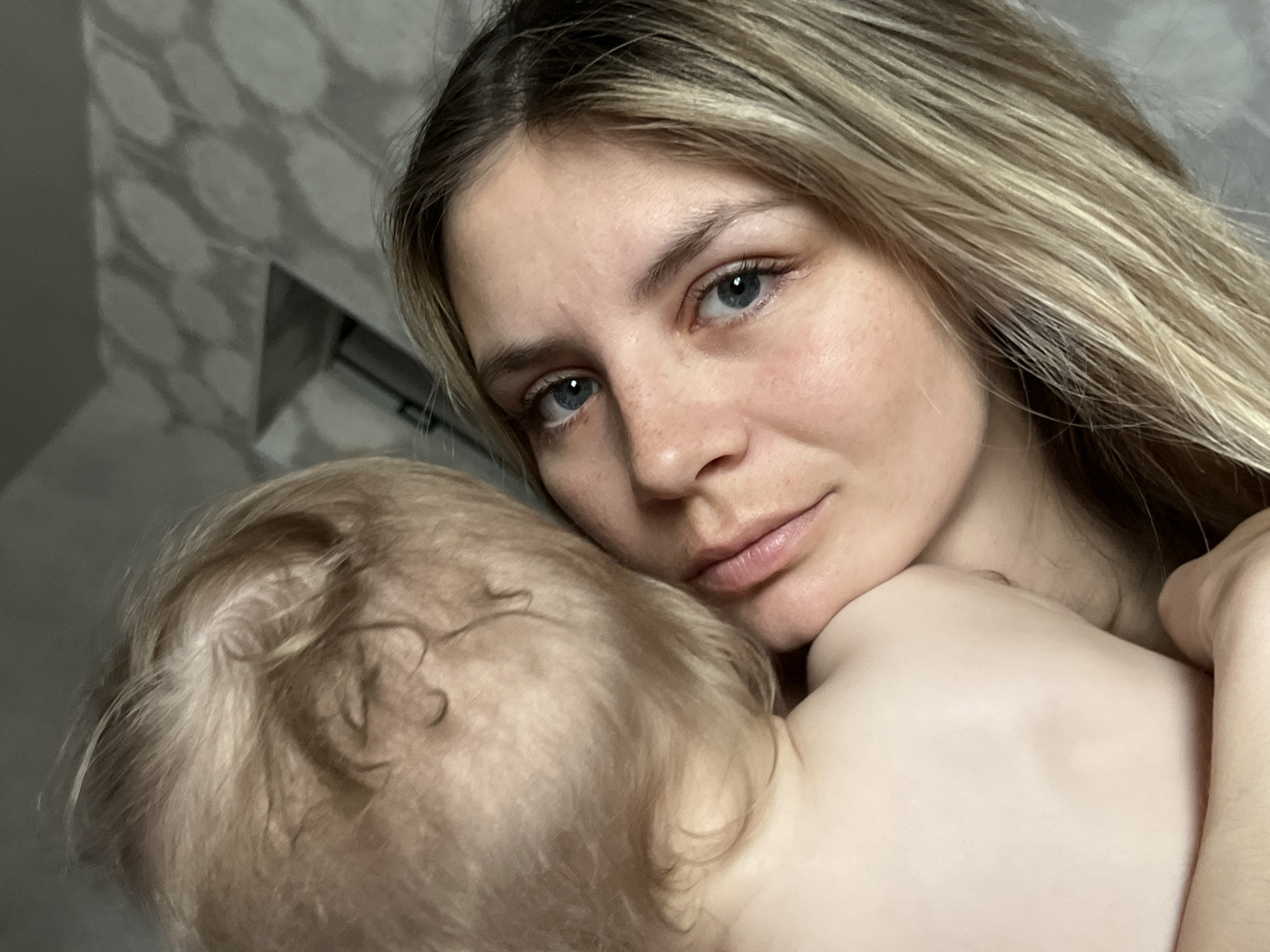A close-up shot shows a fair-skinned woman with blonde hair and blue eyes, holding a fair-skinned child with blonde hair. The woman's face is prominently displayed, while only the back of the child's head is visible. The background is indistinct but appears to be an interior space with patterned wall decor. The location is reported as Chaikovsky, Russia.