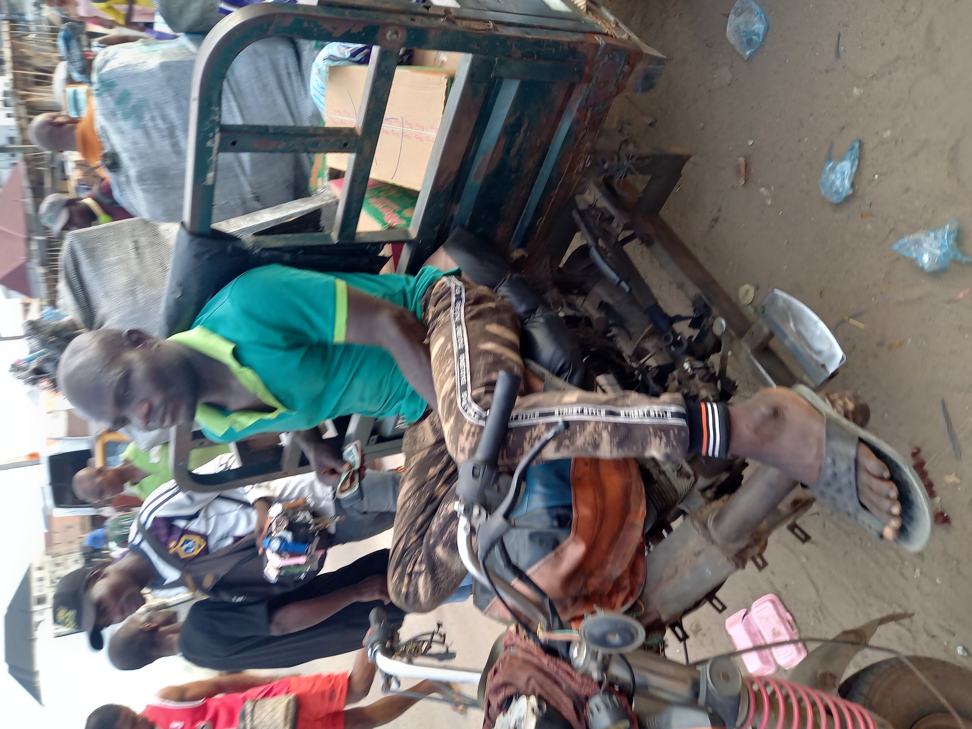 The image shows a scene in what appears to be an outdoor workspace or repair area in Onitsha, Nigeria. A three-wheeled motorcycle (tricycle, commonly known as a "keke") is the central object. The keke has a metal cargo bed and appears to be undergoing repairs, with various mechanical parts visible. Several men are present, some working on or near the keke. One man is wearing a green shirt and patterned pants, while others are dressed in casual clothing. The ground is a mixture of dirt and sand with some scattered plastic bags and debris. The image is taken at a close range, offering a detailed view of the keke and the individuals involved. The surrounding environment appears to be a bustling marketplace or commercial area, with blurred figures in the background.