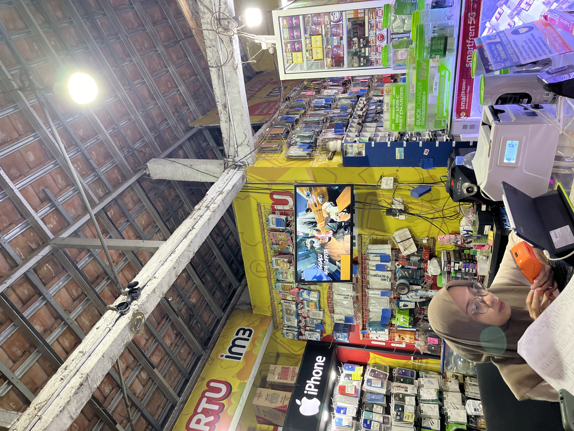 The image captures the interior of a cell phone and accessory shop in Pecangaan Kulon, Indonesia. In the foreground, a woman wearing glasses and a beige hijab is seated at a desk, appearing to work with a portable point-of-sale device and a tablet. She is holding a small orange device in her right hand. The desk is cluttered with papers and other items.
The shop itself is brightly lit and appears to be well-stocked. The walls are adorned with yellow signage, including prominent branding for "IM3" and "iPhone." Numerous shelves are filled with various cell phone cases, chargers, and other accessories, displayed in clear packaging. A television screen mounted on the wall is showing a video of someone playing a guitar.
To the right of the frame, a glass-fronted display cabinet contains rows of cigarette packs, including brands like "Camel," "Black," and "Lucky Strike." Below this, there are shelves displaying fast chargers and other electronic accessories. The ceiling of the shop is visible, featuring exposed rafters and corrugated roofing, indicating a relatively simple construction. The overall environment suggests a busy retail space selling mobile technology and related products.