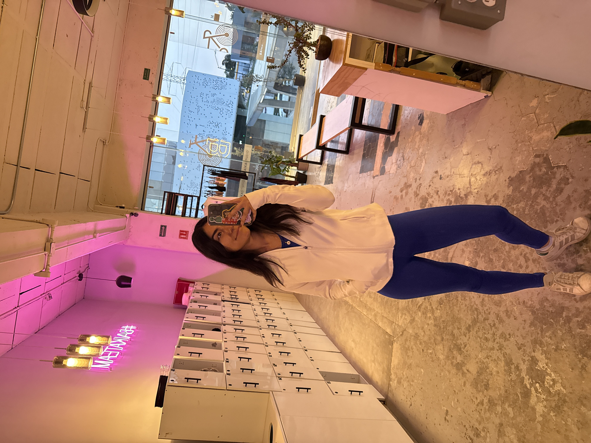 A young woman stands indoors, taking a selfie in what appears to be a mirror or reflective surface. She is wearing a white zipped jacket over a blue top, blue leggings, and white and gray sneakers. Her long dark hair is down, and she holds a smartphone with a decorated case up to her face, showing a slight smile.
The setting is a modern interior space, possibly a gym, studio, or boutique. To her left, a long row of white lockers with black handles extends into the distance, suggesting a changing or storage area. Above the lockers, a neon pink sign glows, displaying the reversed text "#MAESTRANNA". Below the sign, several cylindrical pendant lights hang, emitting a warm, yellowish light. The floor is a textured, light-colored concrete.
To her right, large windows look out onto an urban street scene. Outside, buildings are visible, along with glimpses of power lines. A wooden bench or table is placed by the window, and a small potted plant sits on it. Text visible on structures or signs outside the window includes "BALAN..." and "AIKA STUDIO". An exit sign (green with a white arrow) is mounted on a wall. The overall lighting is a mix of natural light from outside, warm artificial lighting, and the distinct pink glow from the neon sign. The time of day appears to be daylight, possibly late afternoon.