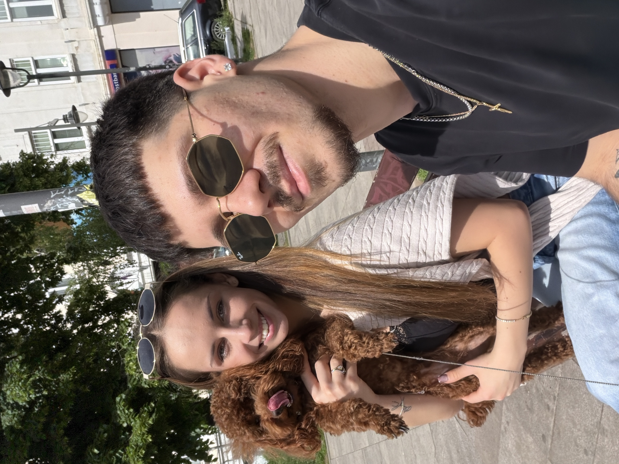 A selfie taken by a couple with their brown poodle in Sofia, Bulgaria. The man is wearing a black t-shirt, gold-framed sunglasses, and a necklace with a cross pendant. He has short dark hair and a beard. The woman is wearing a light-colored knit cardigan and sunglasses perched on her head. She is holding the poodle, which has its tongue sticking out. The background includes a street with trees, buildings, and a parked car. Based on the architectural style and street signage visible, the location is likely in a central area of Sofia.