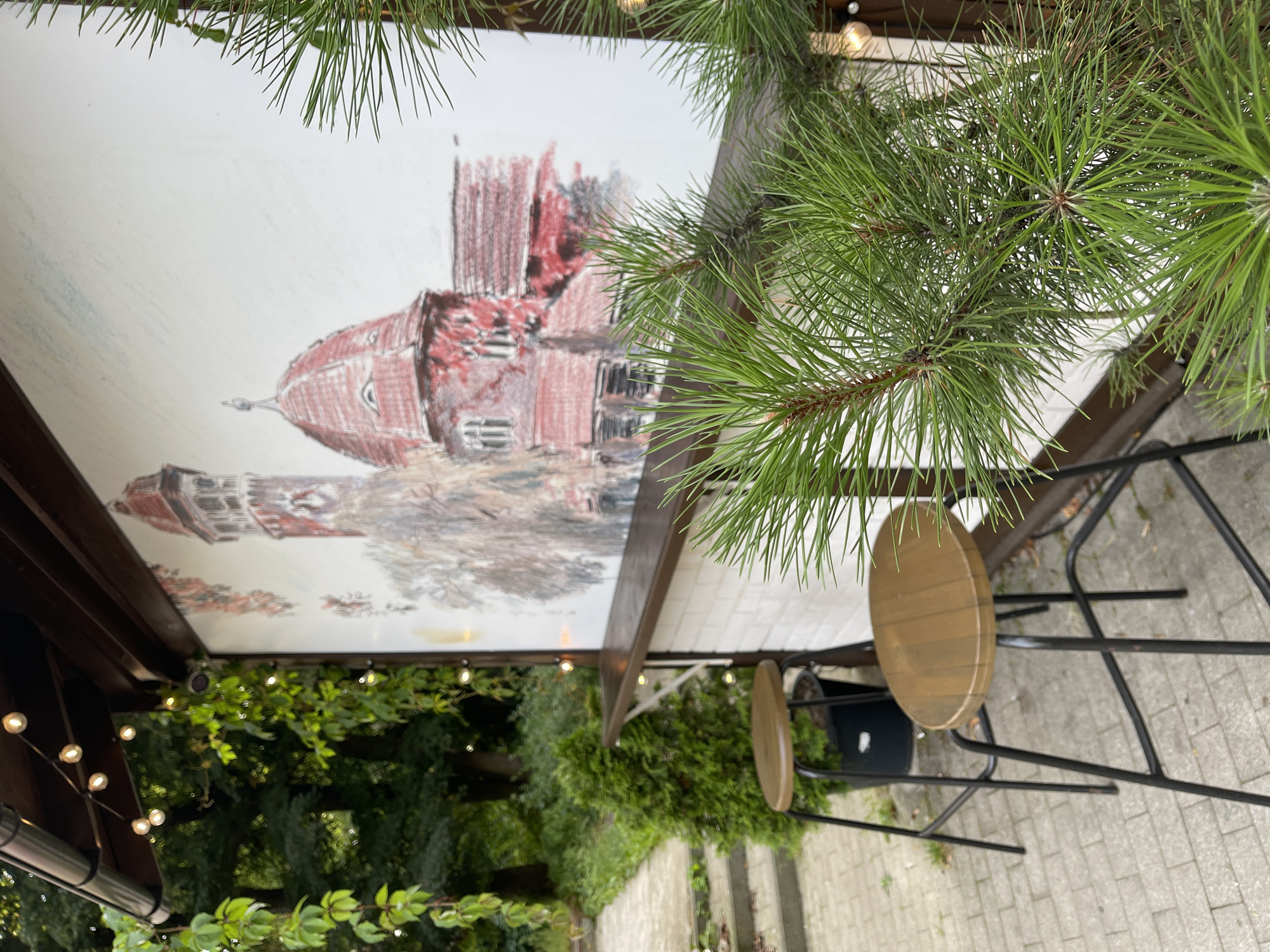 A two-stool outdoor bar area is depicted in Svetlogorsk, Russia, with a mural of a red-roofed building in the background. The bar has a wooden countertop and is backed by a white brick wall. Two wooden-topped bar stools with black metal legs are positioned in front of the bar. A small evergreen tree is planted in a black pot to the left of the bar. To the left of the bar, a paved walkway leads up a set of concrete stairs. Lush green foliage and trees frame the scene. String lights are visible along the edge of the awning above the bar. A pine branch with green needles is in the foreground on the right side of the image.