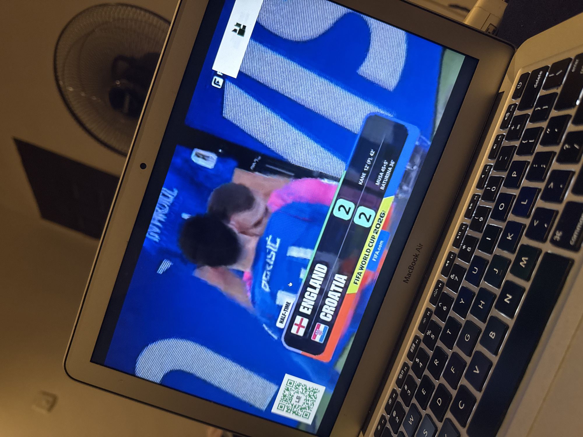 The image shows a laptop screen displaying a soccer match score. On the screen, it shows "HALF-TIME" with England and Croatia tied at 2-2. The game is identified as "FIFA WORLD CUP 2026". Below the score, there are names of players and the times they scored: "KANE 12' (P), 42'", "MUSA 45+5'", and "BATURINA 36'". The laptop is a MacBook Air, and the keyboard is visible in the foreground. A QR code is displayed on the left side of the screen. The background shows a fan and a wall, suggesting an indoor setting. The specific location in Kufena, Nigeria is not discernible from the image.