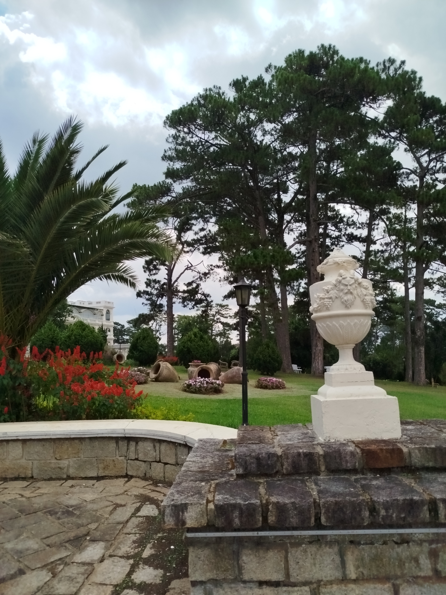 The image depicts a garden setting on an overcast day, seemingly in Da Lat, Vietnam, based on the style of the architecture and landscaping. In the foreground is a stone wall with a white decorative urn on top of it. A stone-paved path leads away from the wall. To the left, there's a raised flower bed with vibrant red flowers and a palm tree. The midground features a well-maintained lawn with several large, overturned earthenware pots arranged as decorative elements. There are also flowerbeds with pink flowers planted inside the pots. A black lamppost stands amongst them. In the background, tall pine trees create a backdrop. A white building with a classical design is partially visible through the trees. The sky is cloudy, suggesting the potential for rain.