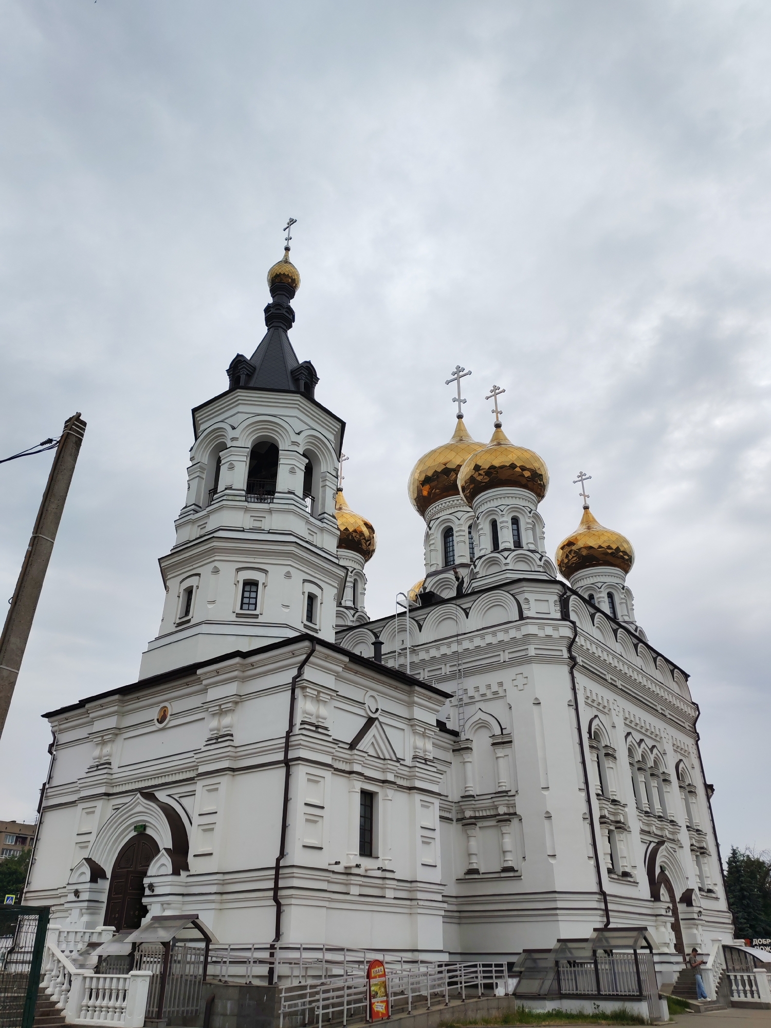 An exterior, low-angle, eye-level shot captures the white facade and golden domes of a large Orthodox church under a cloudy sky. The church features multiple domes topped with crosses. A bell tower with arches and windows is prominent on the left side of the structure. In the foreground, a concrete utility pole stands to the left of the church. White railings and stairs lead up to the main entrance. A ramp is also visible. Several trees and buildings are visible in the background. A person stands on the stairs to the right of the entrance, looking at their phone. Another person is partially visible near the fence. The overall scene is overcast and appears to be in an urban environment.
