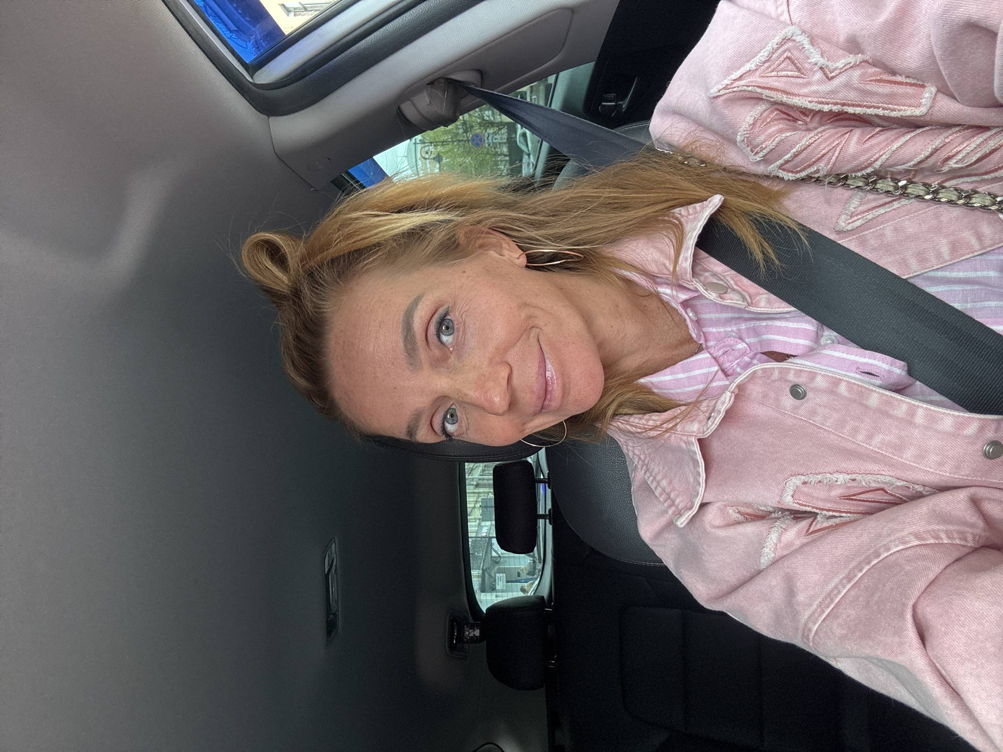 A woman is in a car, wearing a pink jacket and striped pink shirt. She is wearing a seatbelt. The location outside the car is the city of Saint Petersburg, Russia.