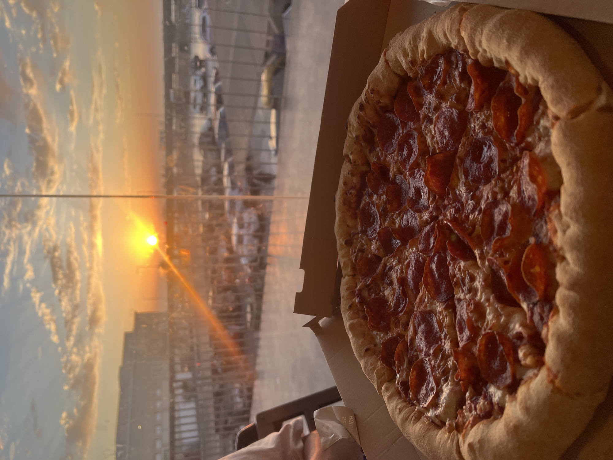 A high-angle shot displays a pepperoni pizza in a cardboard box, situated in front of a window overlooking a sunset in Arica, Chile. The setting sun is low on the horizon, partially obscured by buildings and casting a warm, orange glow across the sky. The window reflections include structures that appear to be part of the city's urban landscape. The pizza is heavily topped with pepperoni and has a thick crust.