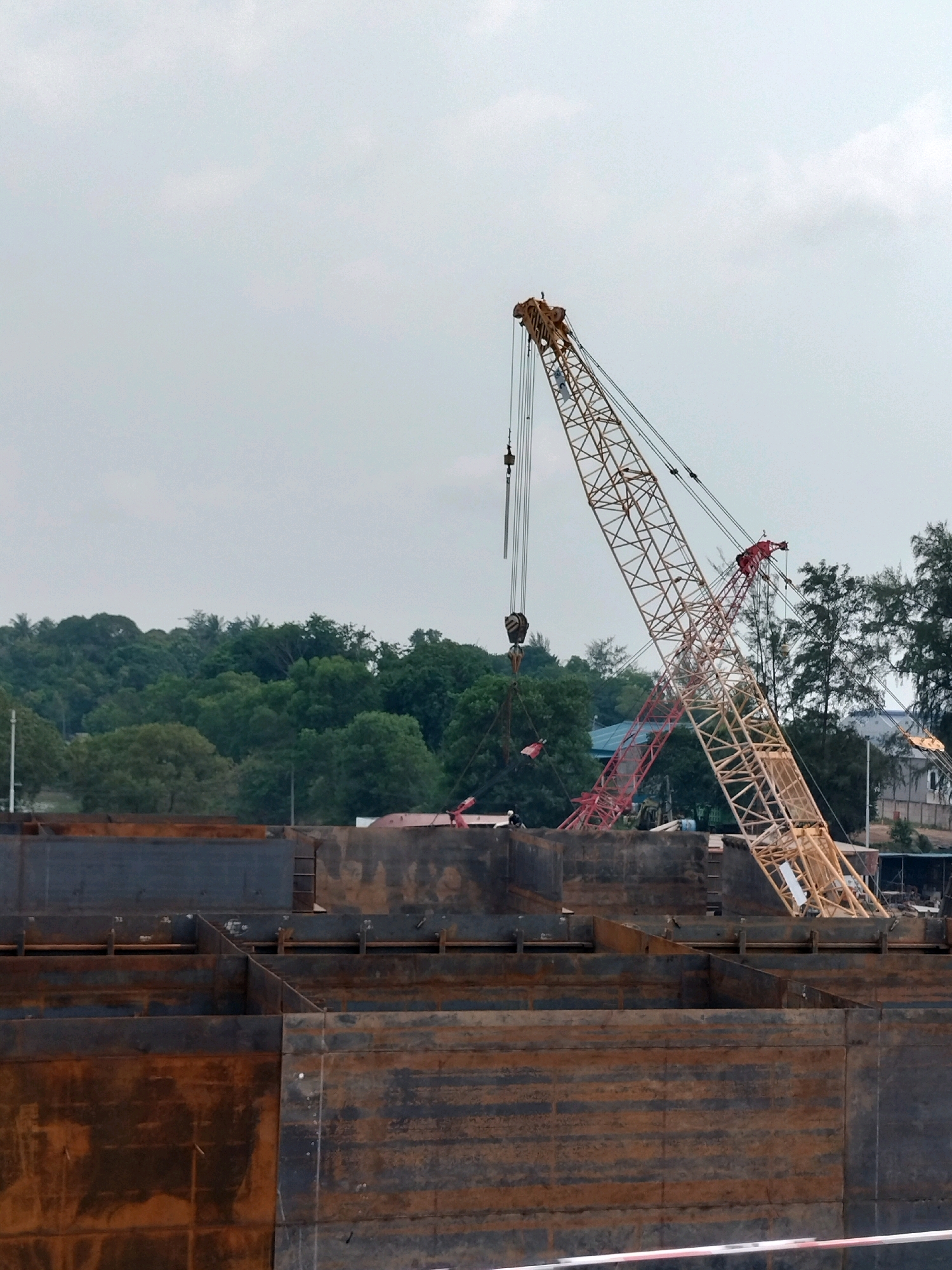 This is an outdoor, daytime, medium shot of a construction site. In the foreground, there are large, rusty metal structures that appear to be part of a ship or barge under construction. Behind these structures, a yellow and red lattice boom crane is prominently visible, with its boom extended upwards and to the right. The crane is suspended by cables. In the background, there is a dense line of lush green trees, suggesting a natural landscape. Partially visible through the trees are some buildings and a lighter colored structure. The sky is overcast with a light grey hue. The scene appears to be located at a shipyard in Batam, Indonesia, a city known for its shipbuilding and industrial activities.