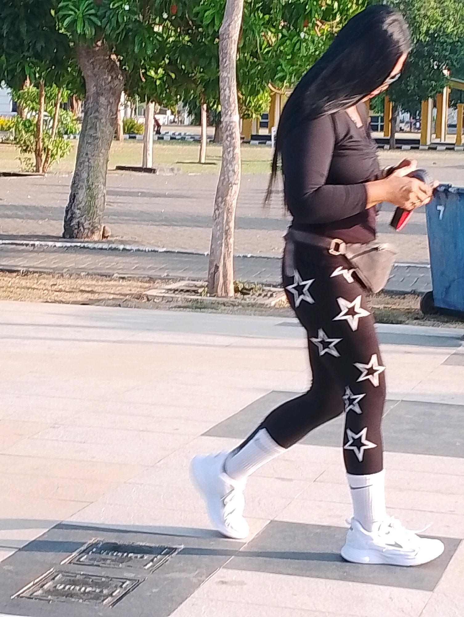 A woman with long, dark hair, wearing a black long-sleeved shirt, black leggings with white stars, white socks, and white sneakers, walks on a paved walkway. She is holding a red object in her right hand. A gray fanny pack is worn around her waist. A large blue trash bin is visible to her right. The background includes trees and a paved road. The scene is likely in a public park or recreational area within the city of Probolinggo, Indonesia, given the presence of lush trees and a generally open, outdoor setting.