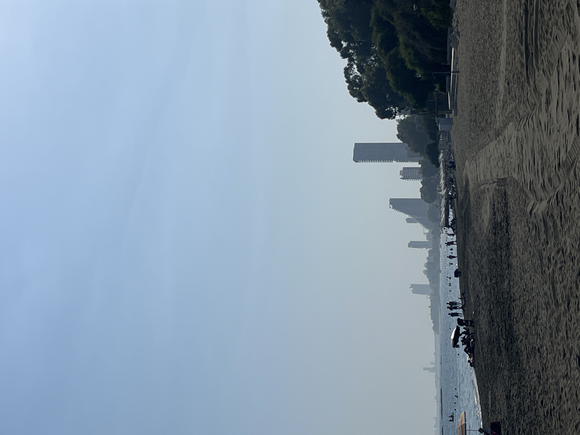 A panoramic view of a sandy beach in Agios Tychona Community, Cyprus, on a clear, hazy day. The foreground is dominated by dark sand with tire tracks from a vehicle, indicating recent activity. In the middle ground, a shoreline separates the beach from the calm sea, where several people are visible swimming and relaxing. A few beach umbrellas and lounge chairs are scattered along the water's edge. Further in the distance, a city skyline emerges, characterized by several modern high-rise buildings, some with distinctive geometric designs, set against a backdrop of lush green trees on a hillside to the right. The sky is a pale blue, fading into a lighter haze near the horizon.