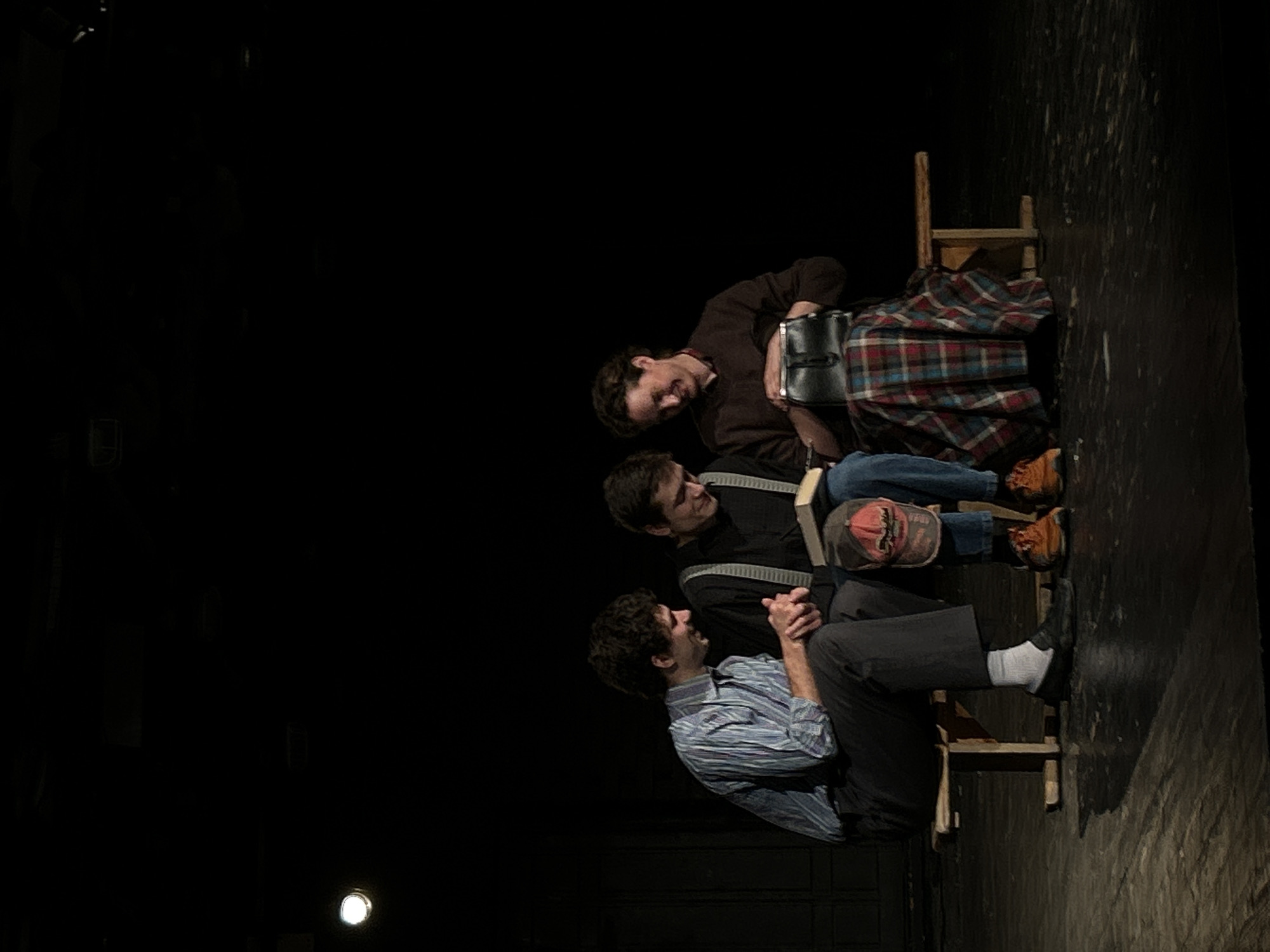 In a dimly lit scene, three individuals are seated on rudimentary wooden chairs. The composition is tilted, with the chairs appearing to be stacked or arranged in a vertical fashion. The background is predominantly black, suggesting a stage or a similarly dark environment.
The person at the top is wearing a dark brown sweater and is looking down and to the right, with a slight smile. They are holding a black bag or satchel. A red, blue, and white plaid fabric is draped over the chair they are sitting on. The middle person is wearing a dark shirt and straps across their chest, and is looking towards the person below them with an intense expression. They are holding a book with a light-colored cover. The bottom person, wearing a blue and white striped shirt, is looking up and to their right, their hands clasped in their lap, conveying a sense of engagement or contemplation. Their legs are bent at the knees, with one foot resting on a rung of the chair. They are wearing dark pants and white socks, with black shoes.
The scene appears to be a theatrical performance or rehearsal, given the stage lighting and the dramatic arrangement of the figures. The atmosphere is somber, with the focus entirely on the individuals and their interactions. There is no visible text in the image.