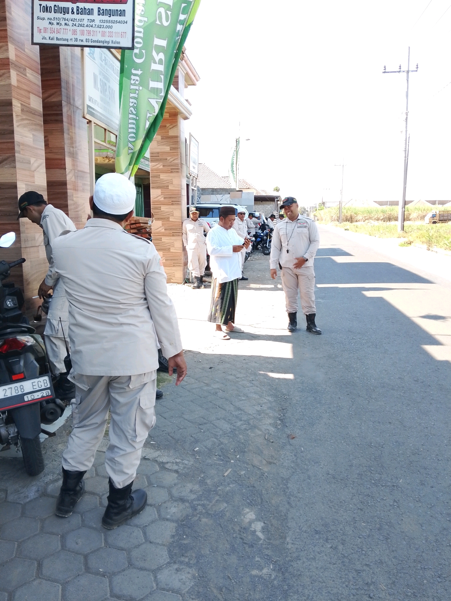 A man wearing a white cap and light-colored uniform stands with his back to the camera, facing a street in Banjarejo, Indonesia. He has black boots and his pants have cargo pockets. To his left, another man in a similar uniform and black cap is leaning forward, looking down. In the background, several other people are visible, some in light-colored uniforms and one in a white shirt and sarong. A motorcycle is parked on the left side of the image, with a license plate reading "2788 ECB 10.28". A tall utility pole stands on the right side of the street. The scene appears to be outdoors on a sunny day, with a paved road and a sidewalk made of hexagonal tiles. A green banner with text is partially visible in the upper left of the image, and a building with a brown tiled facade is next to the sidewalk.