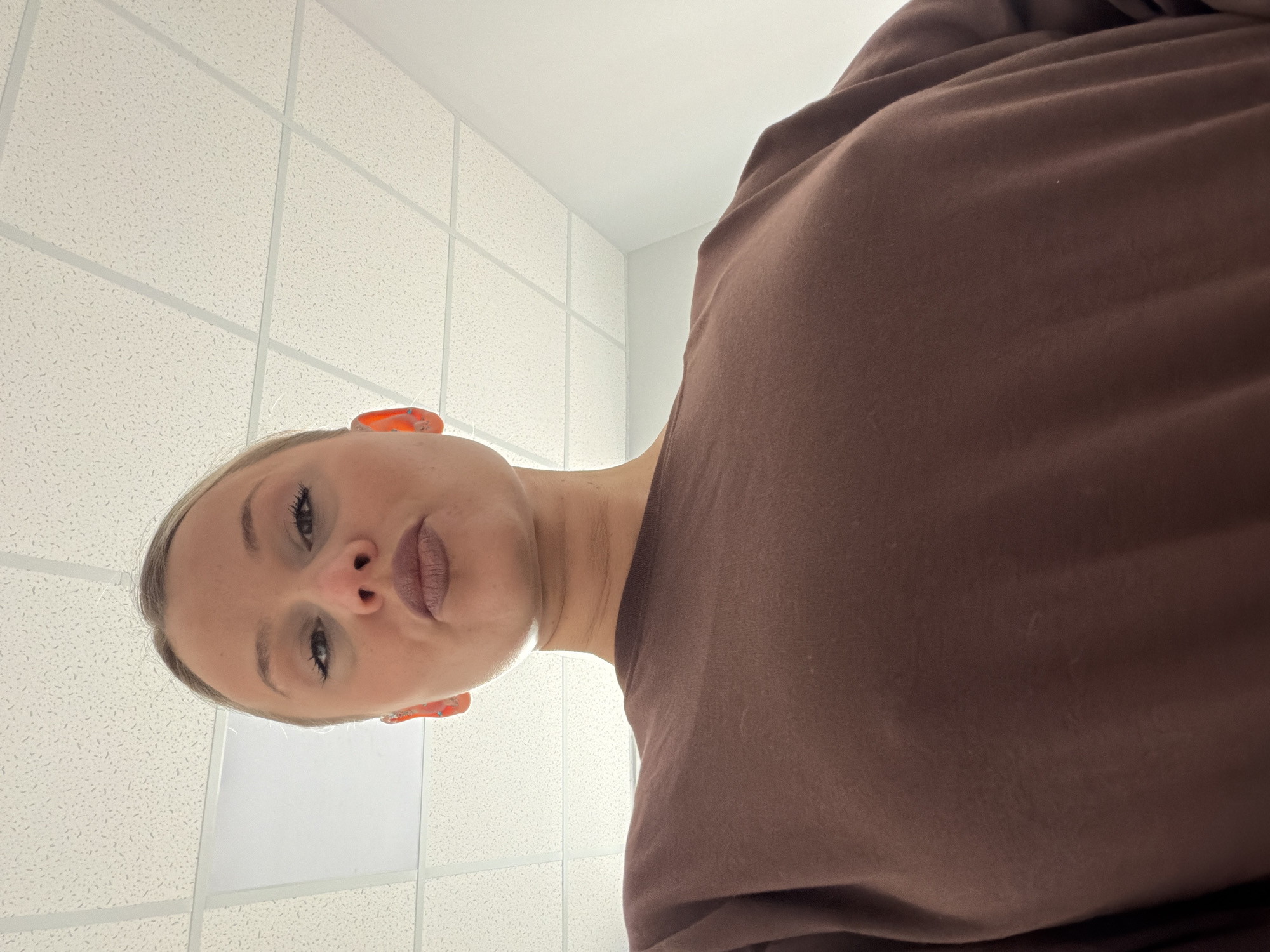A low-angle shot looking up at a light-skinned person with blond hair and light eyebrows. The person is wearing a brown sweater. The background features a white tiled ceiling and a white wall. The location is not identifiable as Sarapul, Russia.
