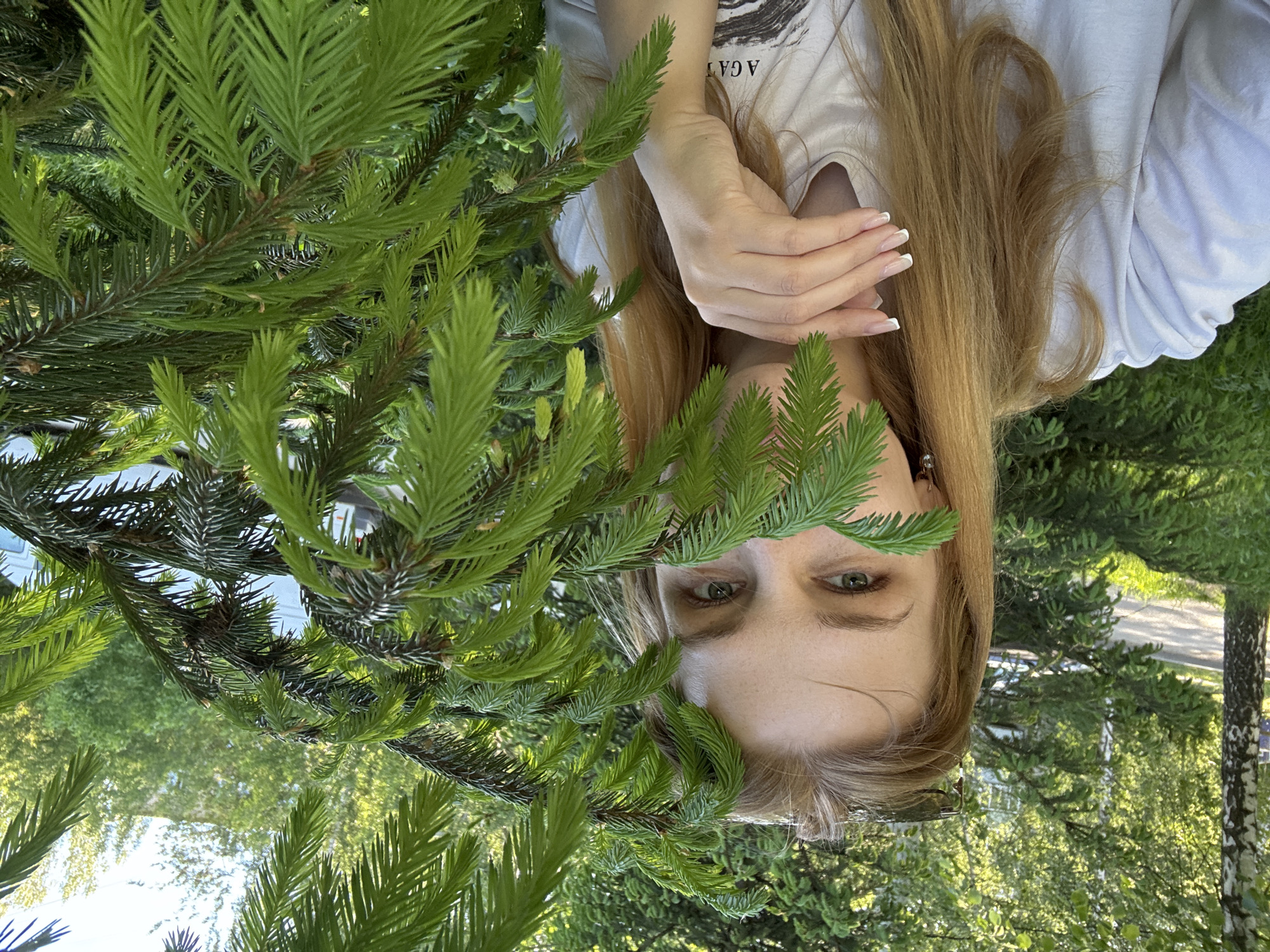 A young woman with long, light brown hair is positioned upside down behind the foliage of a coniferous tree, obscuring most of her face, except for her eyes and forehead. Her left hand is visible near her face, showcasing French-manicured nails. She is wearing a white T-shirt with partial writing on it. The location appears to be outdoors, potentially in a park or garden, indicated by the surrounding trees and greenery. Based on the provided city name (Rusanovo, Russia), the image likely depicts a local resident engaging in leisure or a photographic activity within a green space in the city. Other trees and a pole are visible in the background.