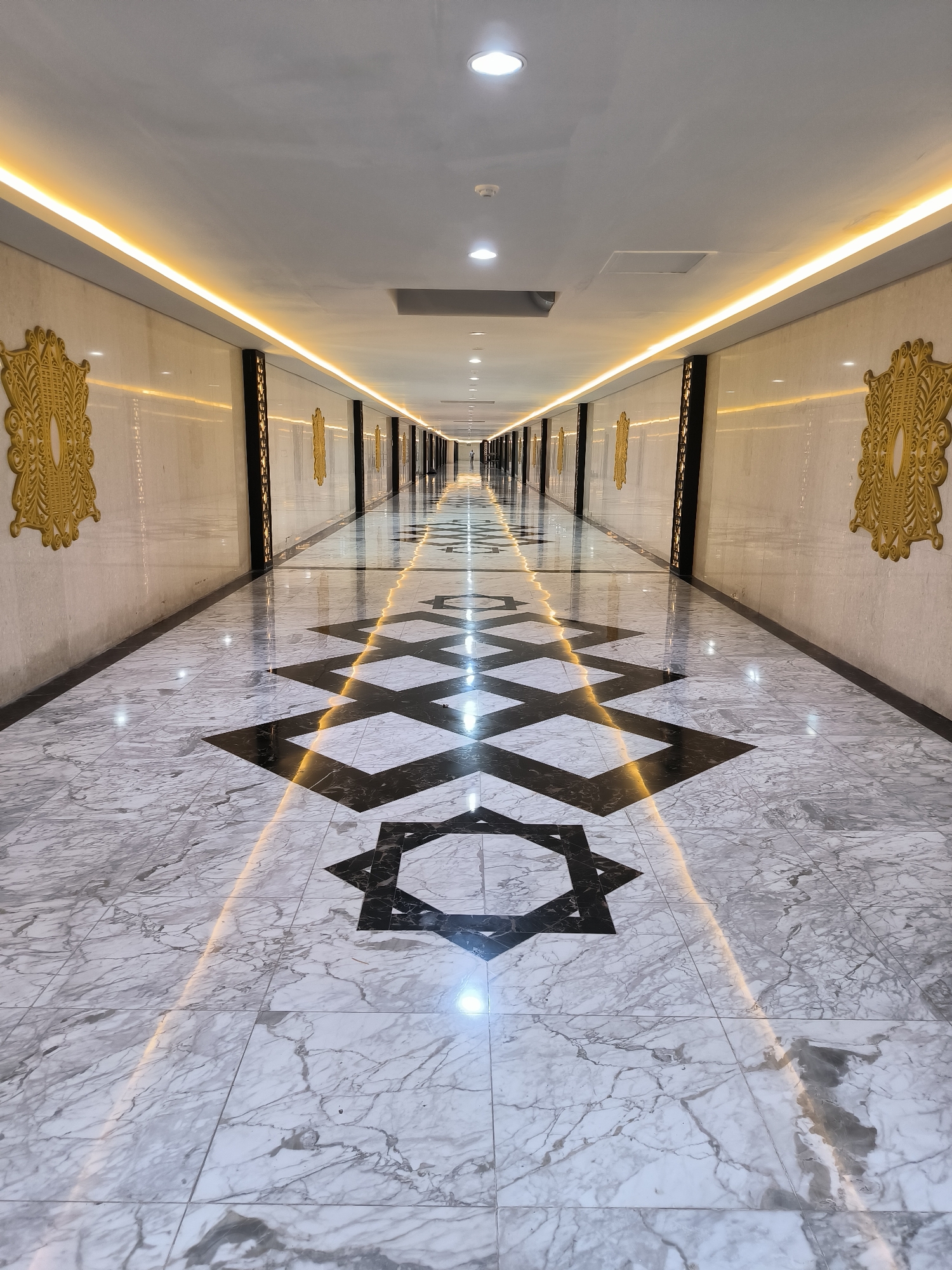 This image shows a long, wide, modern hallway with a high ceiling. The walls are made of polished marble and feature decorative golden crests at intervals. The floor is also marble with a geometric black pattern and is illuminated by a warm, yellow light strip running along the center. Recessed lighting fixtures are visible on the ceiling, along with a ventilation grate. The hallway recedes into the distance, creating a strong sense of perspective. At the far end of the hallway, a person can be seen standing. The scene appears to be an interior space, possibly a hotel, convention center, or a similar public building. Based on the question, this location is identified as being in Banda Aceh, Indonesia.