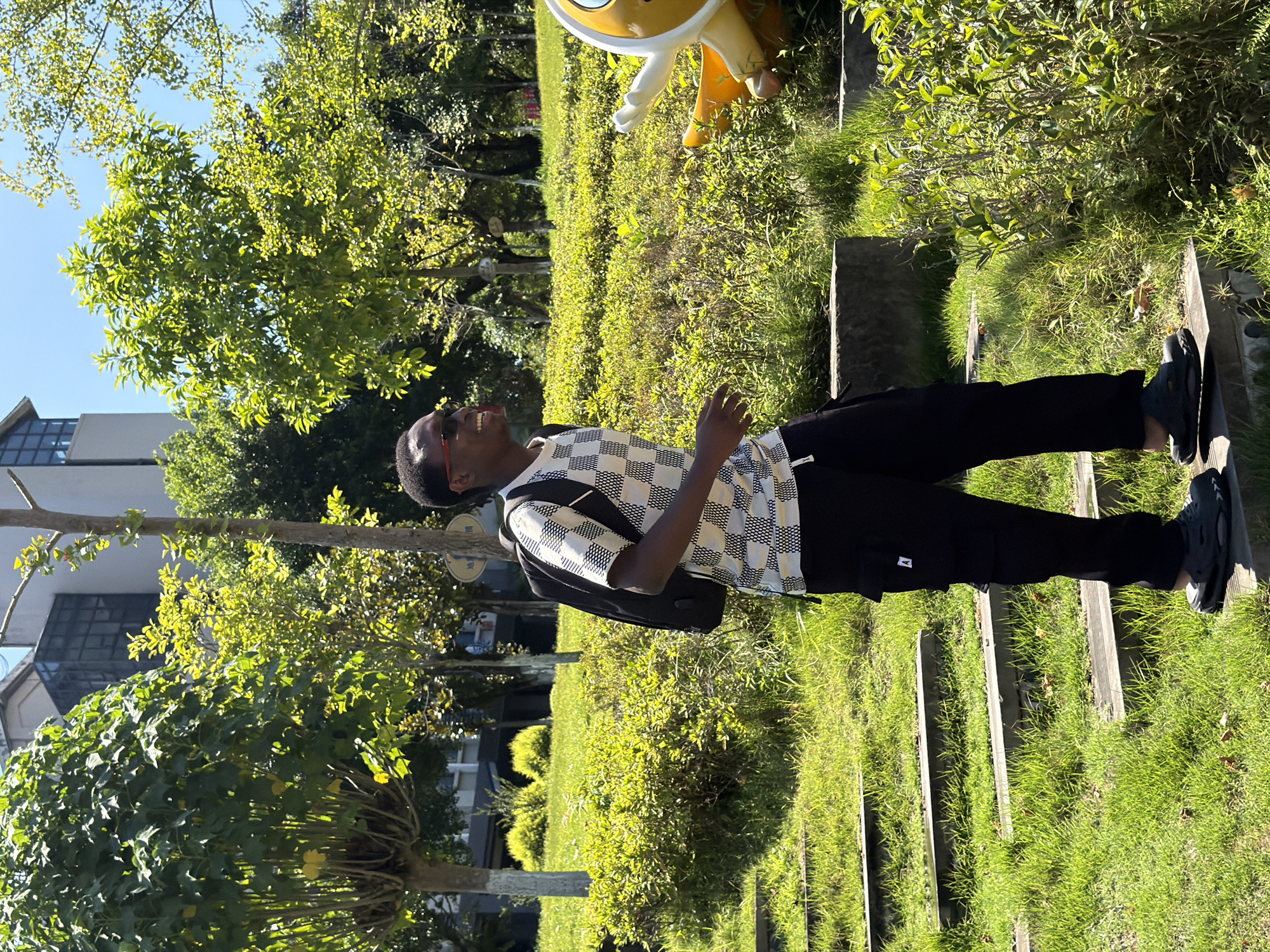 A young Black man with dark skin and short, black hair is walking up a set of outdoor stairs in a park on a sunny day. He is wearing red-rimmed sunglasses, a short-sleeved shirt with a black and white checkered pattern, black cargo pants, and black slip-on sandals. He has a black backpack on his shoulders. The stairs are made of wood and stone and are covered in green grass. The park is lush with green trees and bushes. In the background, there is a modern building with glass windows. To the right of the man, there is a large, yellow, cartoon-like statue with white accents. The scene appears to be in a public park in the city of Jiangjin, China, given the context of the request.
