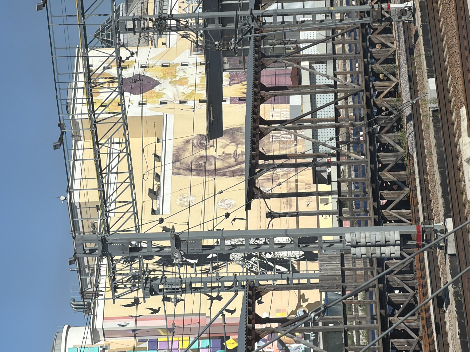 The image shows a mural on the side of a building, as seen from a railway track. The mural features a large portrait of a person's face, rendered in a sepia tone. To the left of the face, there are silhouette images of birds in flight, painted in black. The building itself is painted in shades of yellow and beige, with some sections having a striped pattern of pastel colors. Above the mural, there are visible ventilation pipes and lighting fixtures. In the foreground, there is a wooden pedestrian bridge supported by metal and wood structures, with a railway track running beneath it. The sky is clear and blue. The mural is located on the side of the Dynamo Stadium building in Moscow, Russia.
