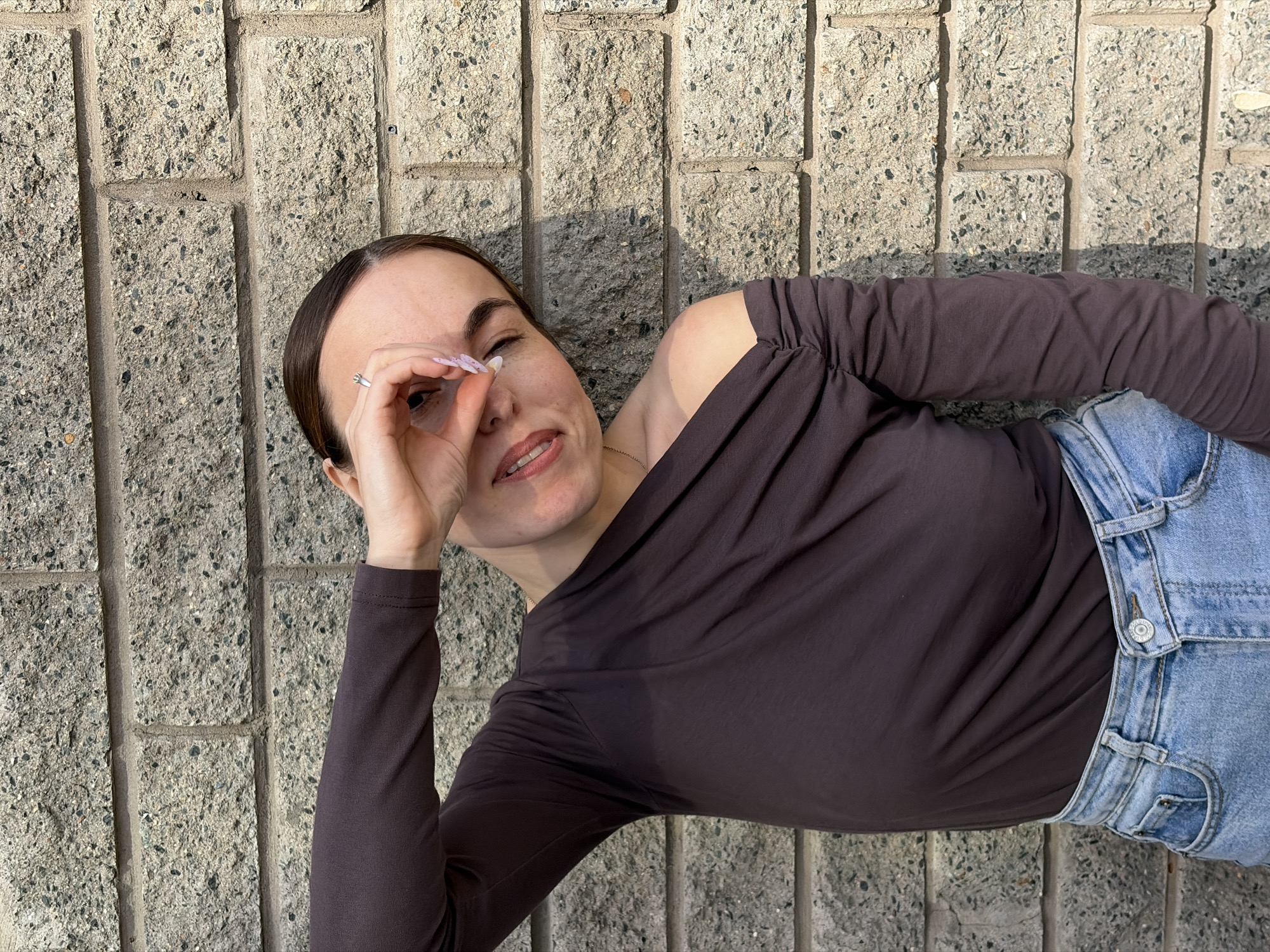 A woman with light skin and dark hair is laying on a light-colored concrete brick surface. She is wearing a dark brown long-sleeved shirt and blue denim jeans. Her left hand is raised to her left eye, forming a circle with her fingers. She is wearing a ring on her left ring finger and her nails are painted a light color. The background is a textured concrete brick wall. The lighting suggests it is daytime. No information is present to suggest this photo was taken in Izhevsk, Russia.