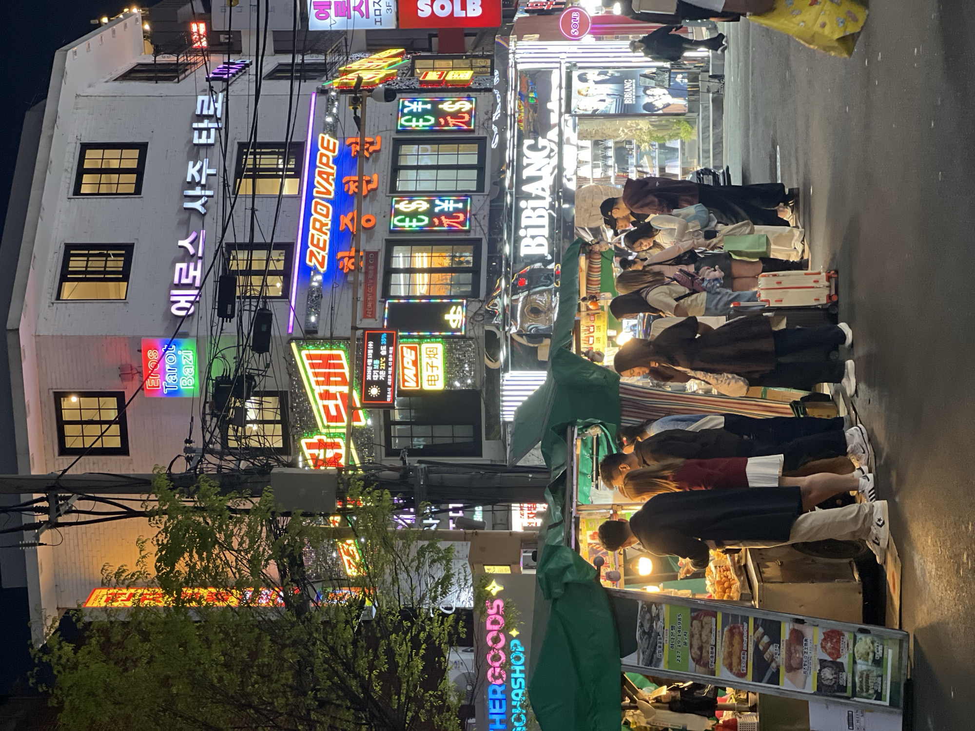 This image shows a nighttime street scene in Seoul, South Korea. People stand in line at food stalls, with several displaying menus featuring various Korean street foods. Buildings in the background are illuminated with neon signs in Korean and English, advertising businesses such as "ZERO VAPE," "BIBIANG," and "Eros Tarot Bazi." Overhanging power lines are visible against the buildings. A tree with illuminated branches stands in the foreground, partially obscuring the view.
