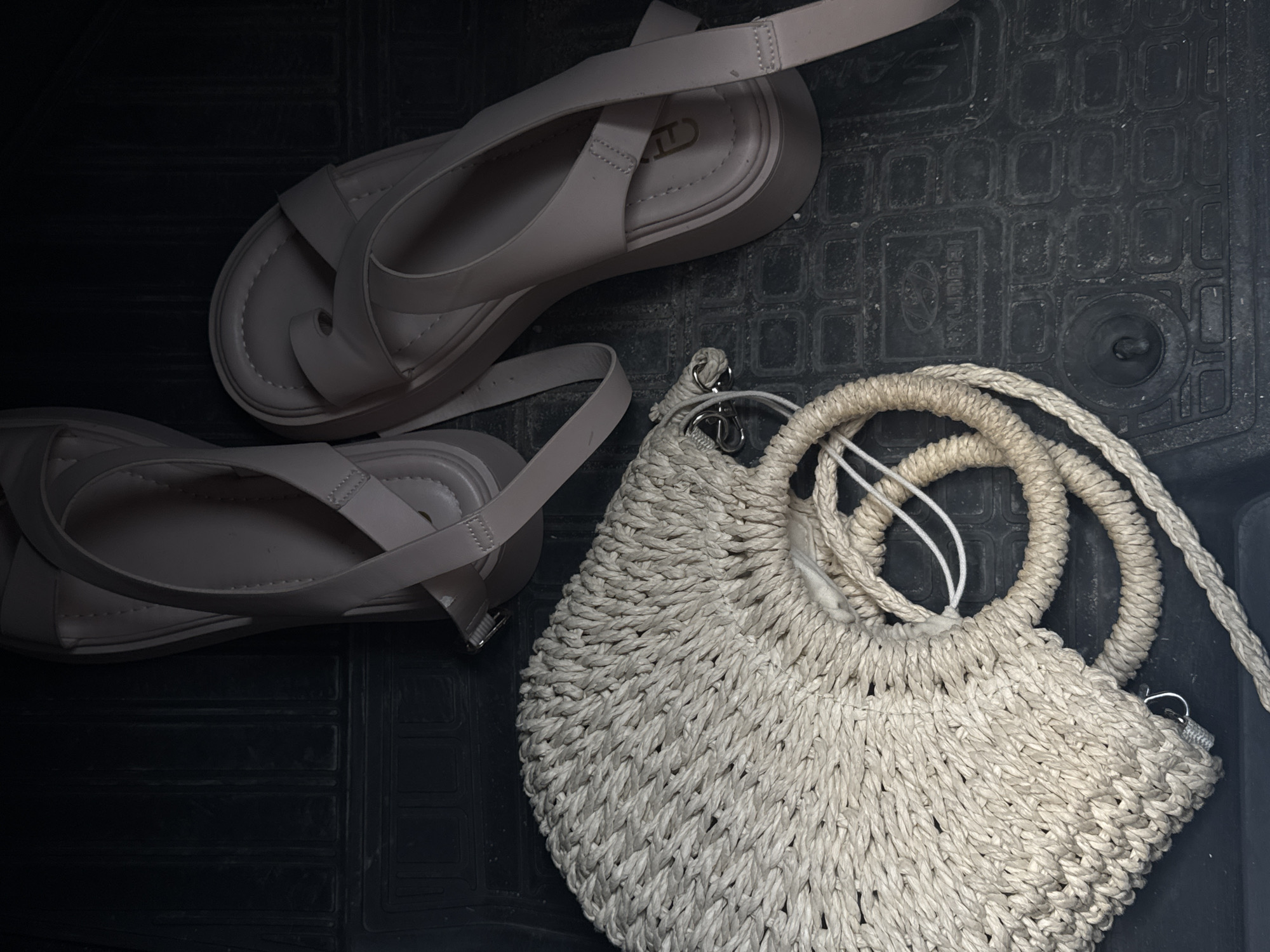 The image shows a pair of light-colored sandals and a white woven bag on a dark, textured surface. The sandals are arranged on the left side of the frame, one slightly above the other. The bag is on the right side. The location is Unknown, Unknown.