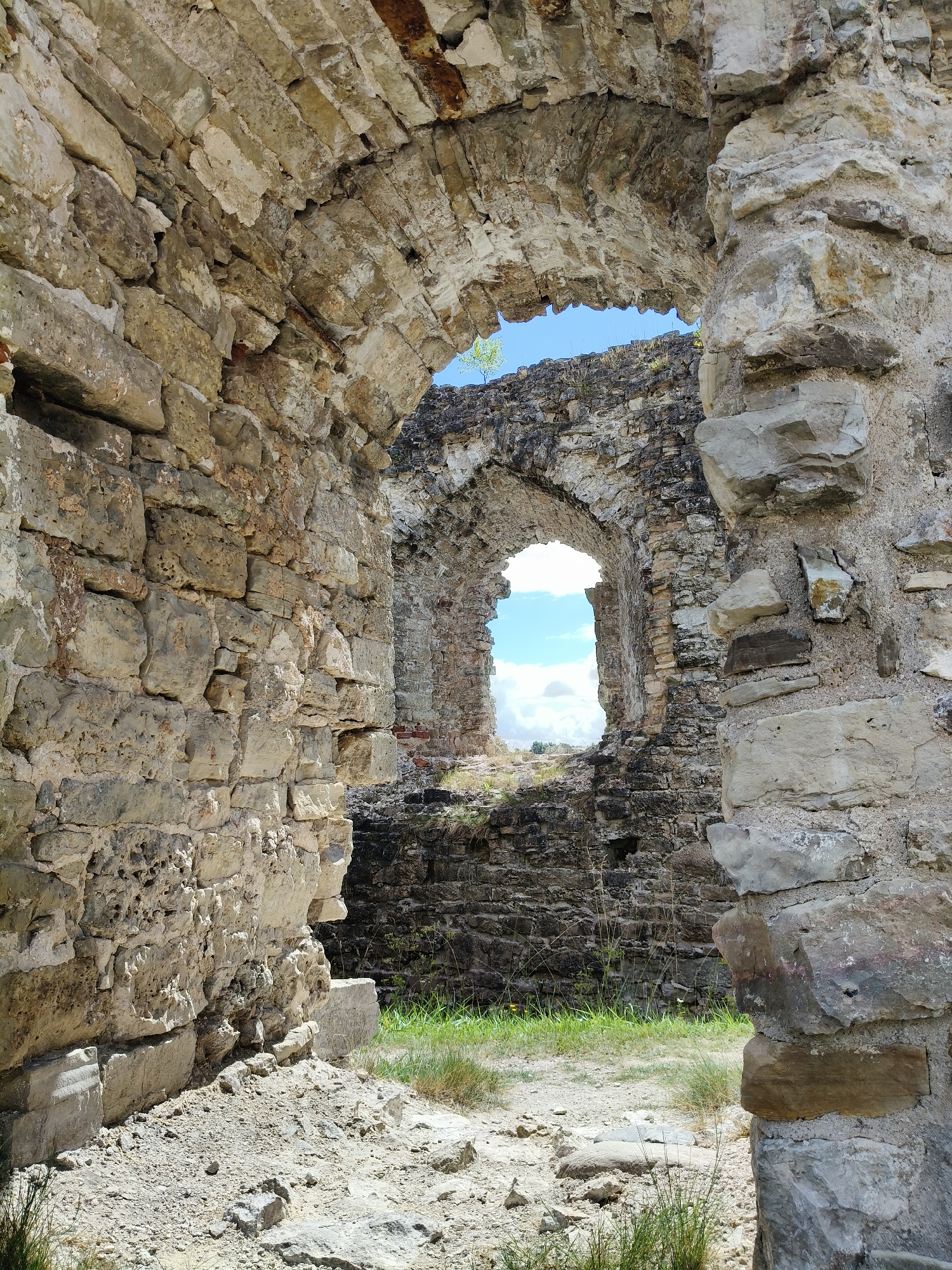 The image shows the ruins of the Livonian Order Castle in Koknese, Latvia. The foreground features a crumbling stone archway, with rough-hewn stones forming the structure. Beyond the arch, another, smaller arched opening reveals a view of the sky and clouds. The walls are made of large, irregular stones, some of which are partially covered in lichen or moss. The ground in front of the ruins is covered with gravel and patches of dry grass. The overall impression is one of ancient decay and enduring history.
